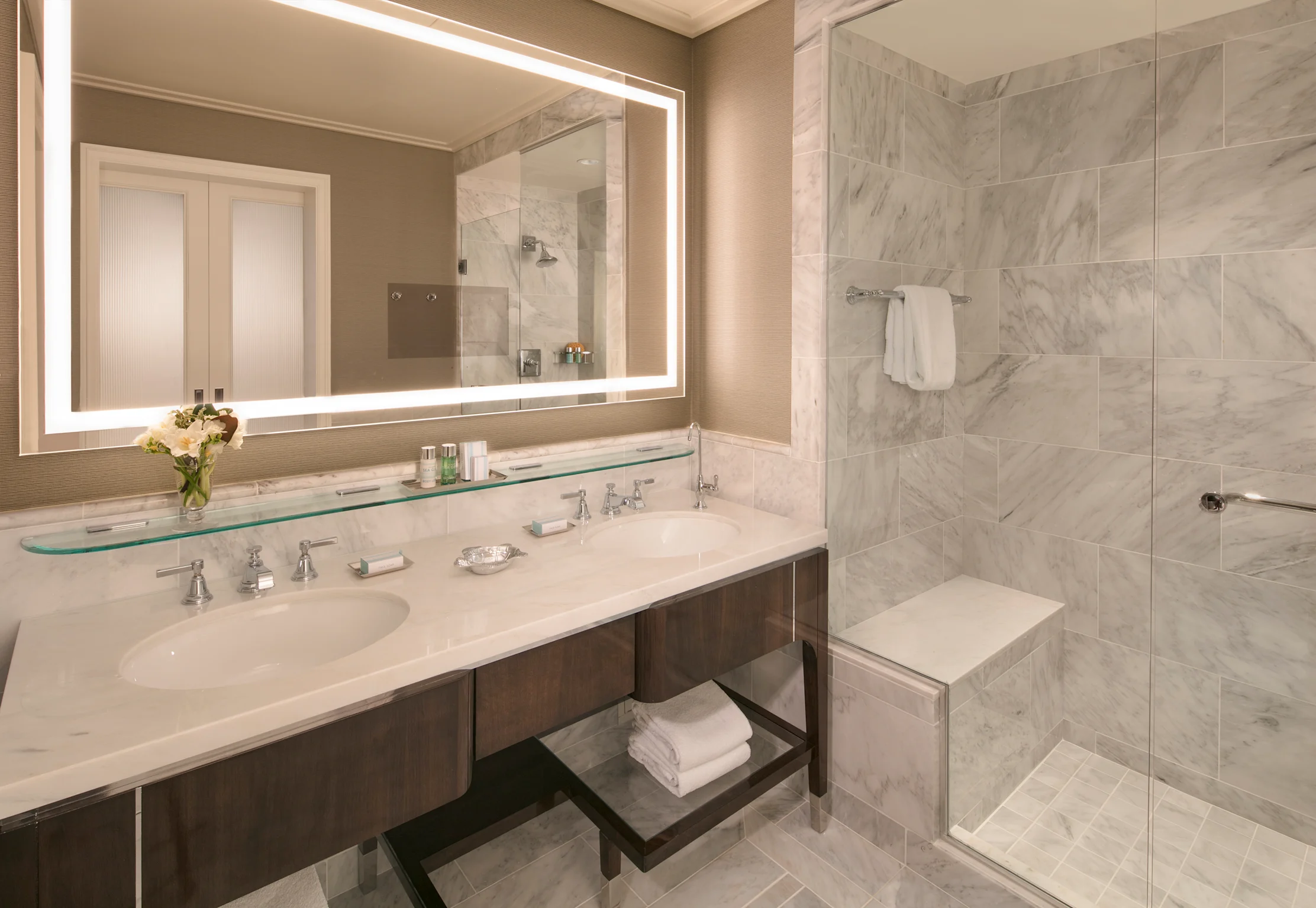 Modern bathroom with marble tiles, backlit mirror, double vanity, glass shower, and white towels.