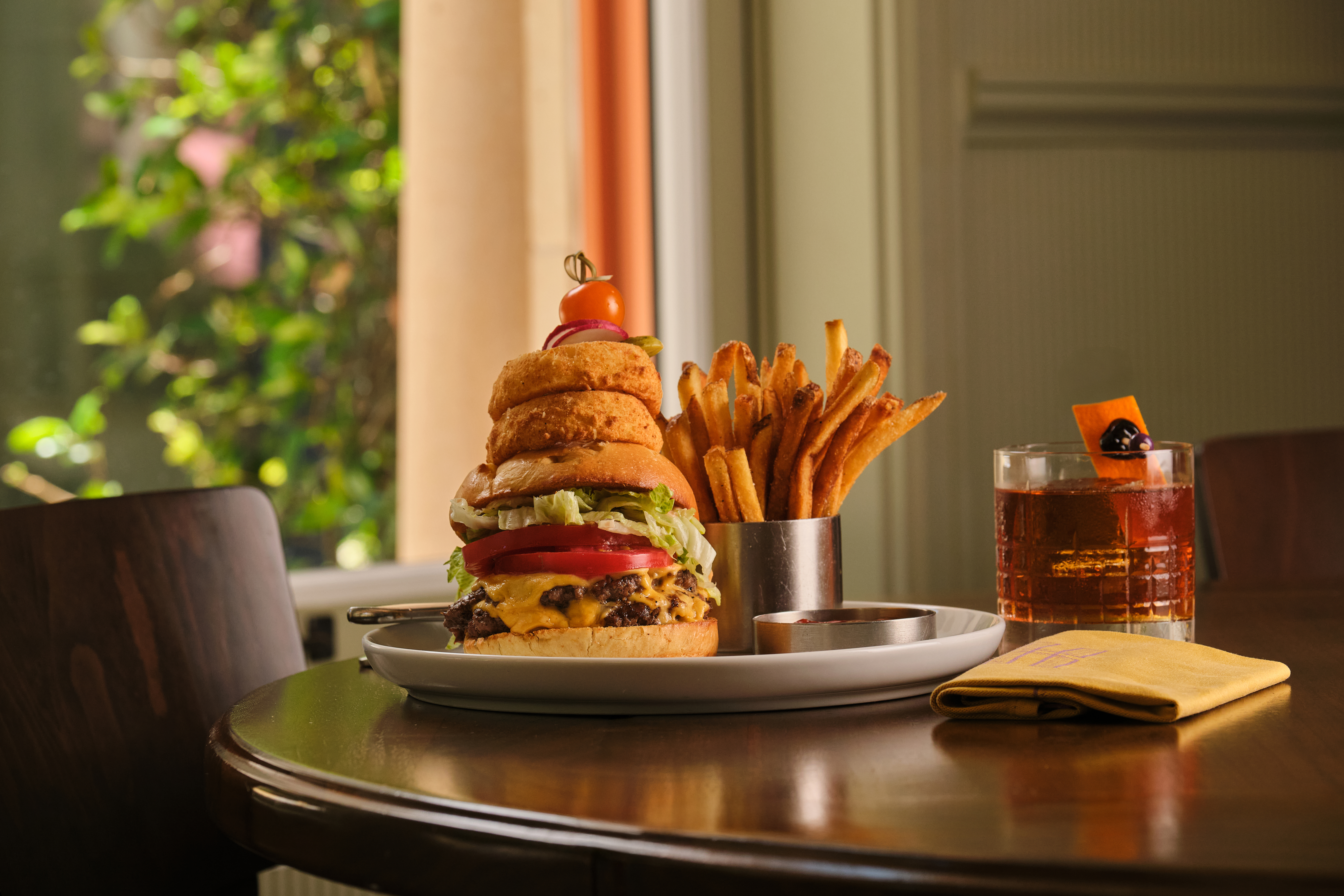Stacked burger with onion rings, fries in metal cup and cocktail on table near window.