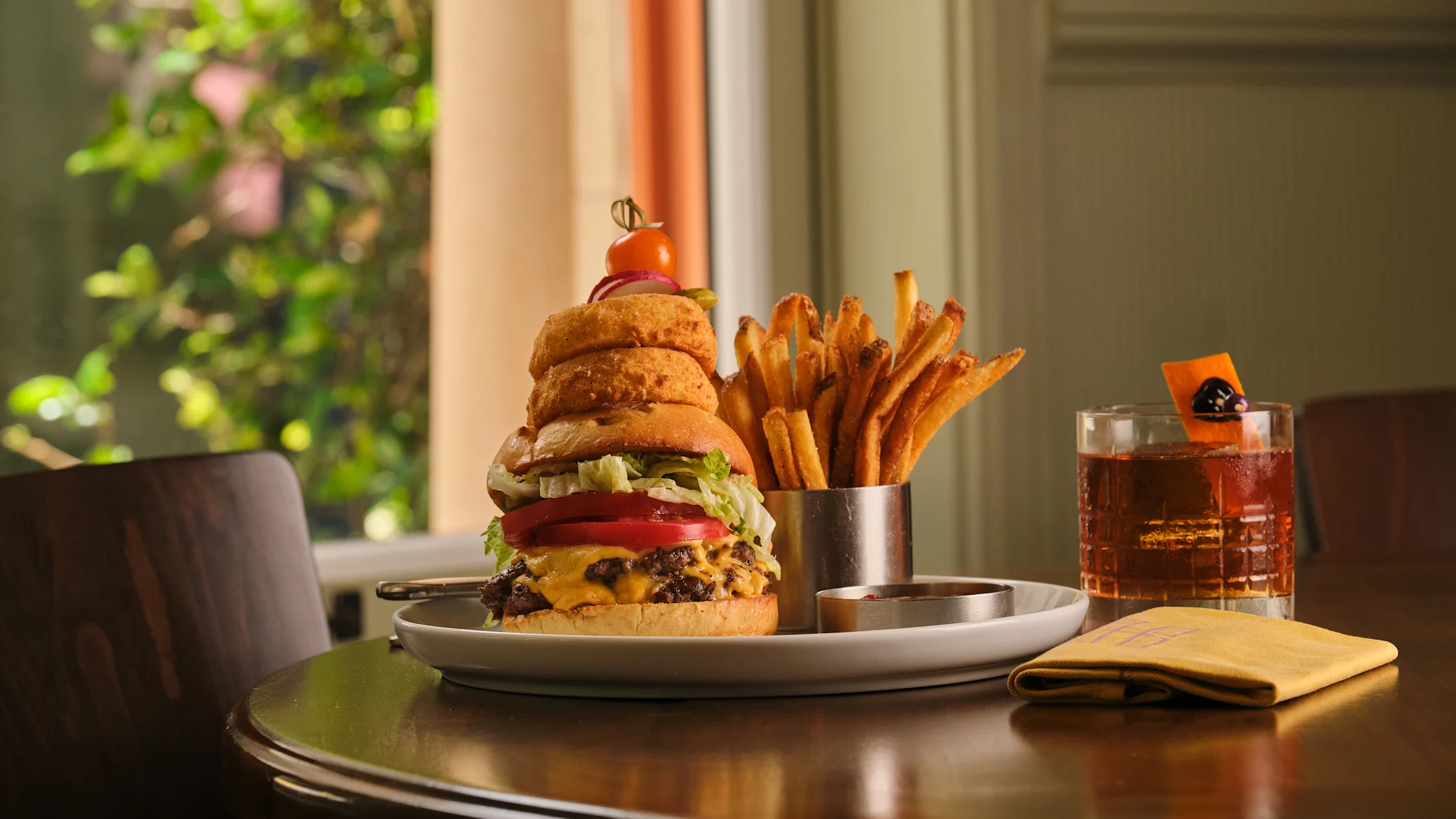 Stacked burger with onion rings, fries in metal cup and cocktail on table near window.