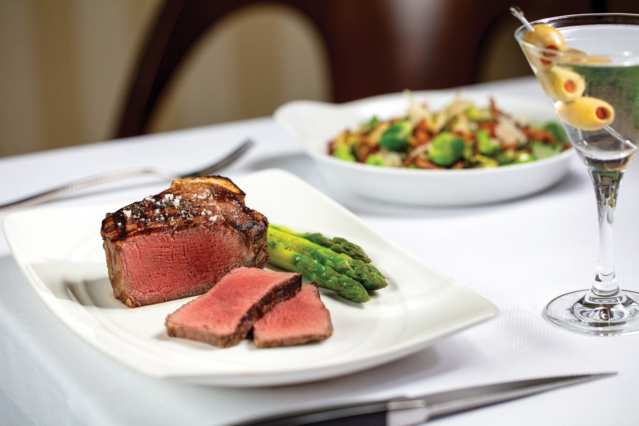 Grilled steak with asparagus on white plate, salad bowl, and martini with olives on table.