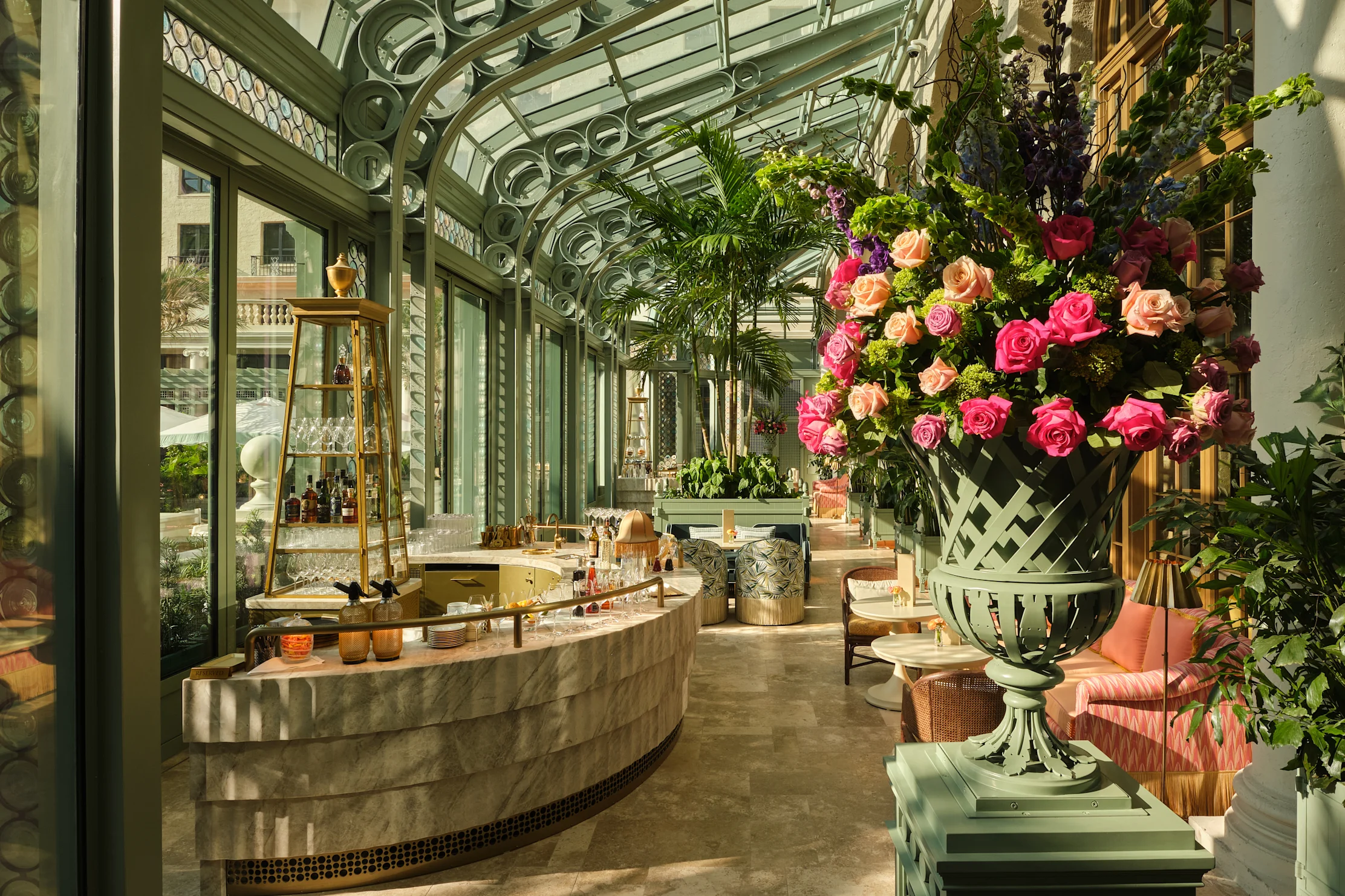 Elegant conservatory lounge with green glass ceiling, tropical plants, marble bar, and coral seating