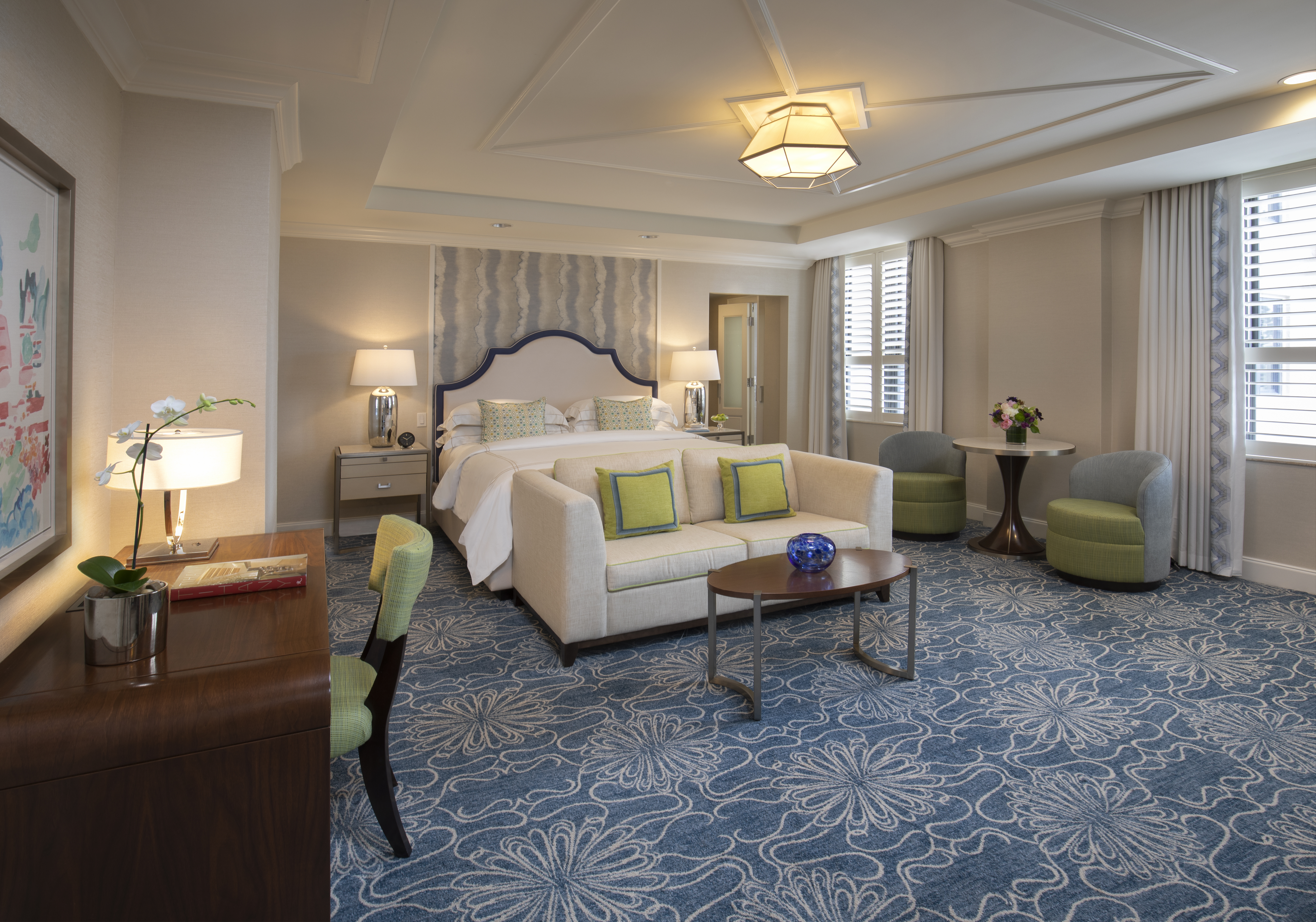 Elegant hotel suite with beige sofa, green accent pillows, bed with upholstered headboard, and blue patterned carpet.