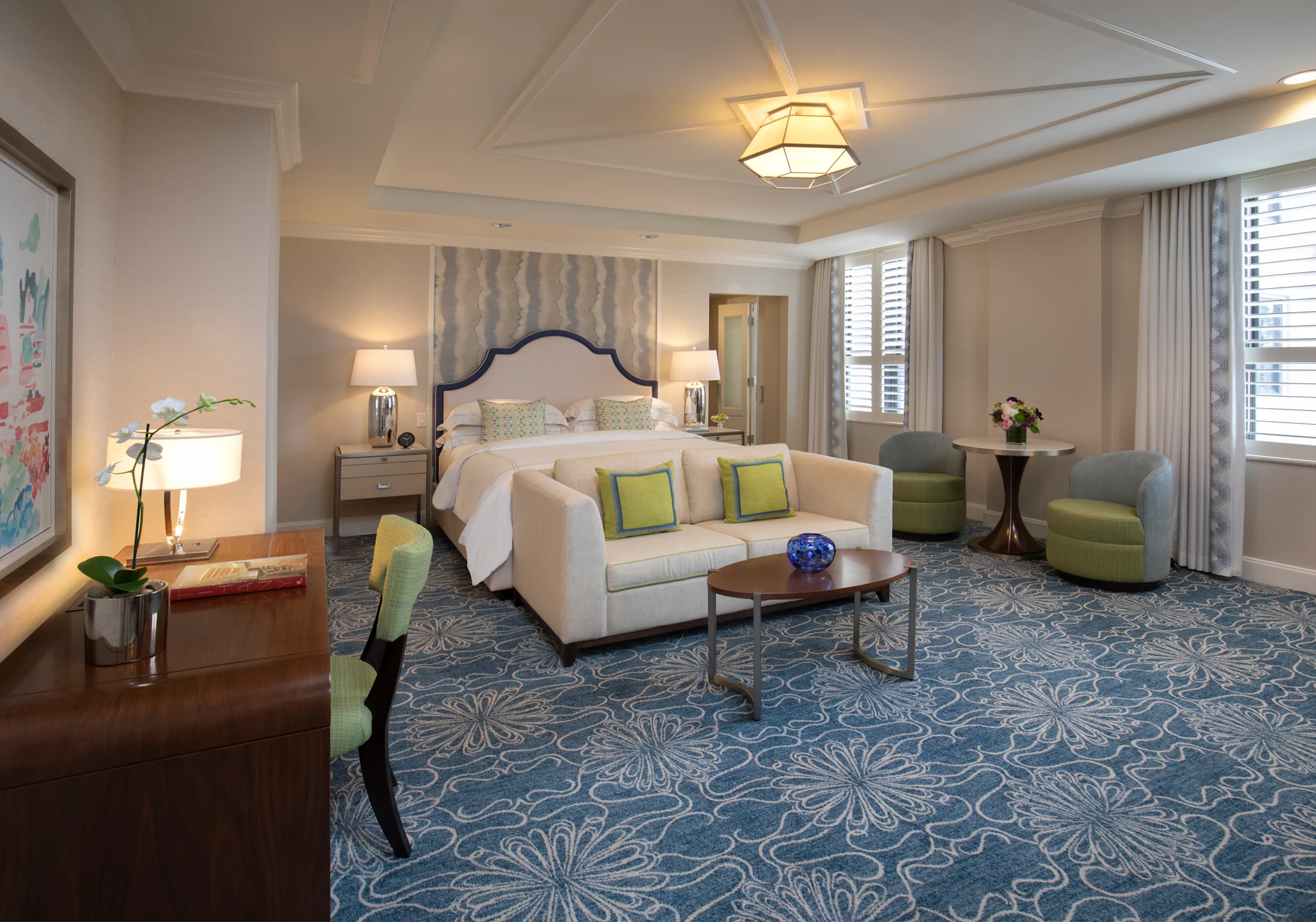 Elegant hotel suite with beige sofa, green accent pillows, bed with upholstered headboard, and blue patterned carpet.