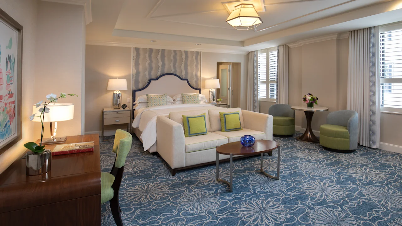 Elegant hotel suite with beige sofa, green accent pillows, bed with upholstered headboard, and blue patterned carpet.
