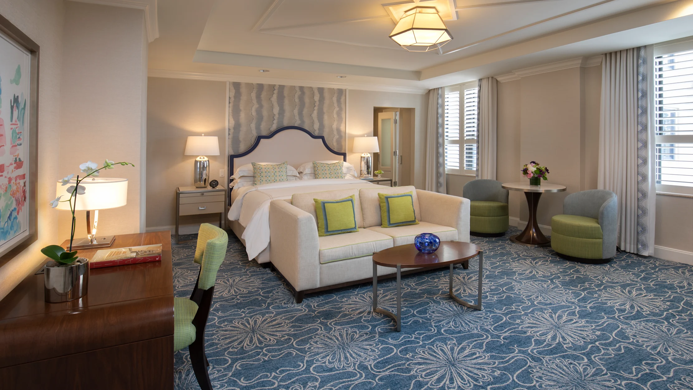 Elegant hotel suite with beige sofa, green accent pillows, bed with upholstered headboard, and blue patterned carpet.