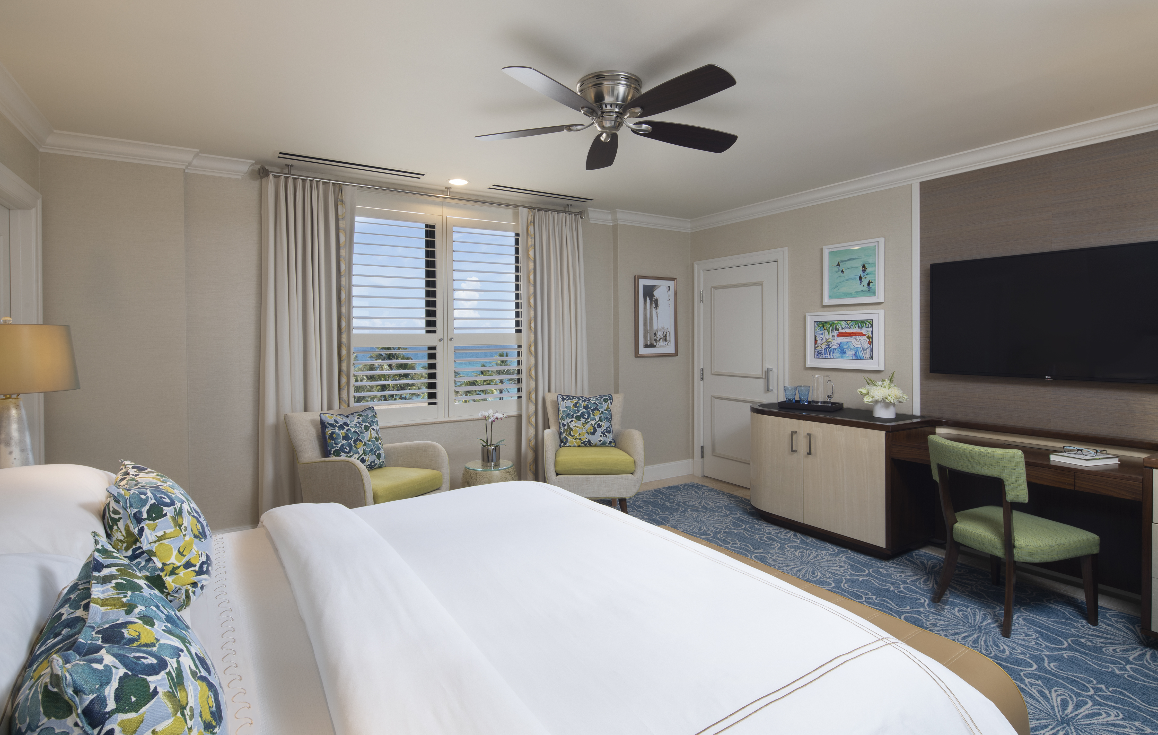 Bright hotel room with white bed, blue patterned pillows, ceiling fan, ocean view windows, and TV.