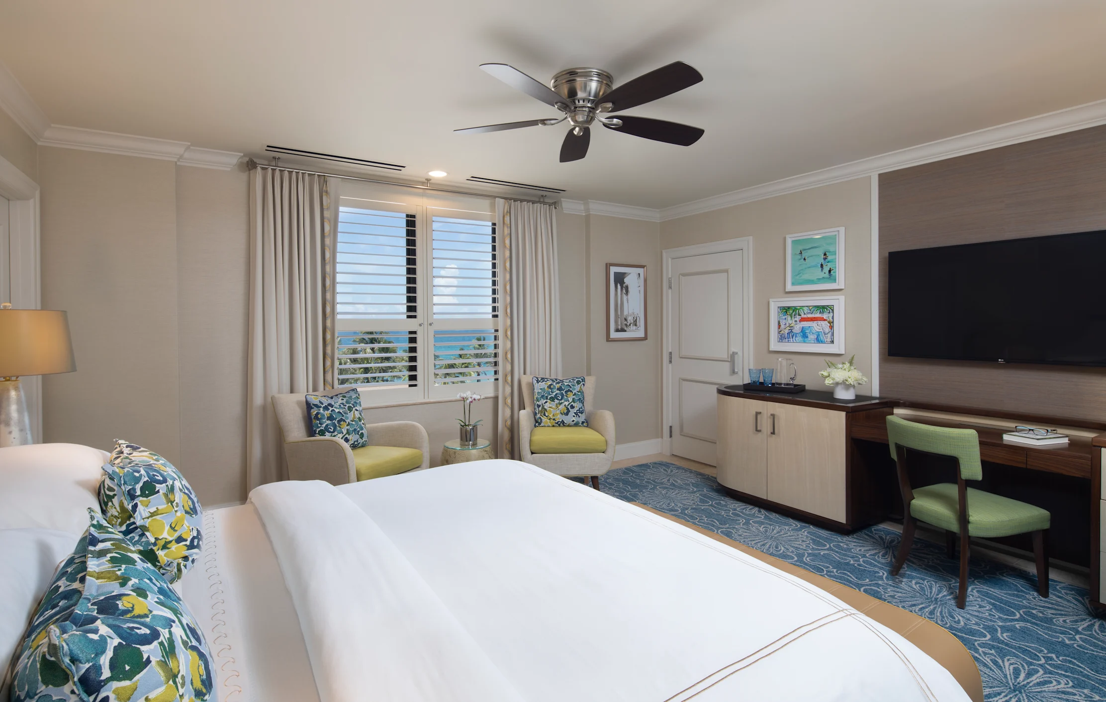 Bright hotel room with white bed, blue patterned pillows, ceiling fan, ocean view windows, and TV.