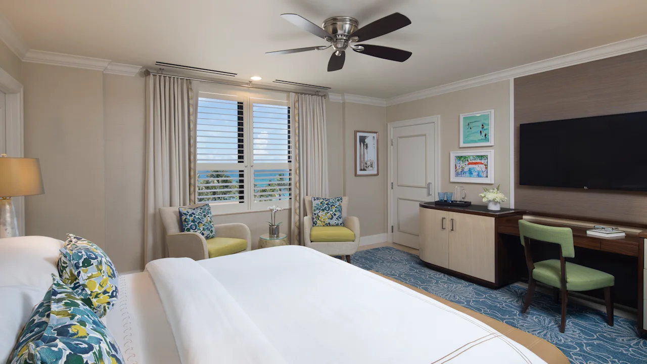Bright hotel room with white bed, blue patterned pillows, ceiling fan, ocean view windows, and TV.