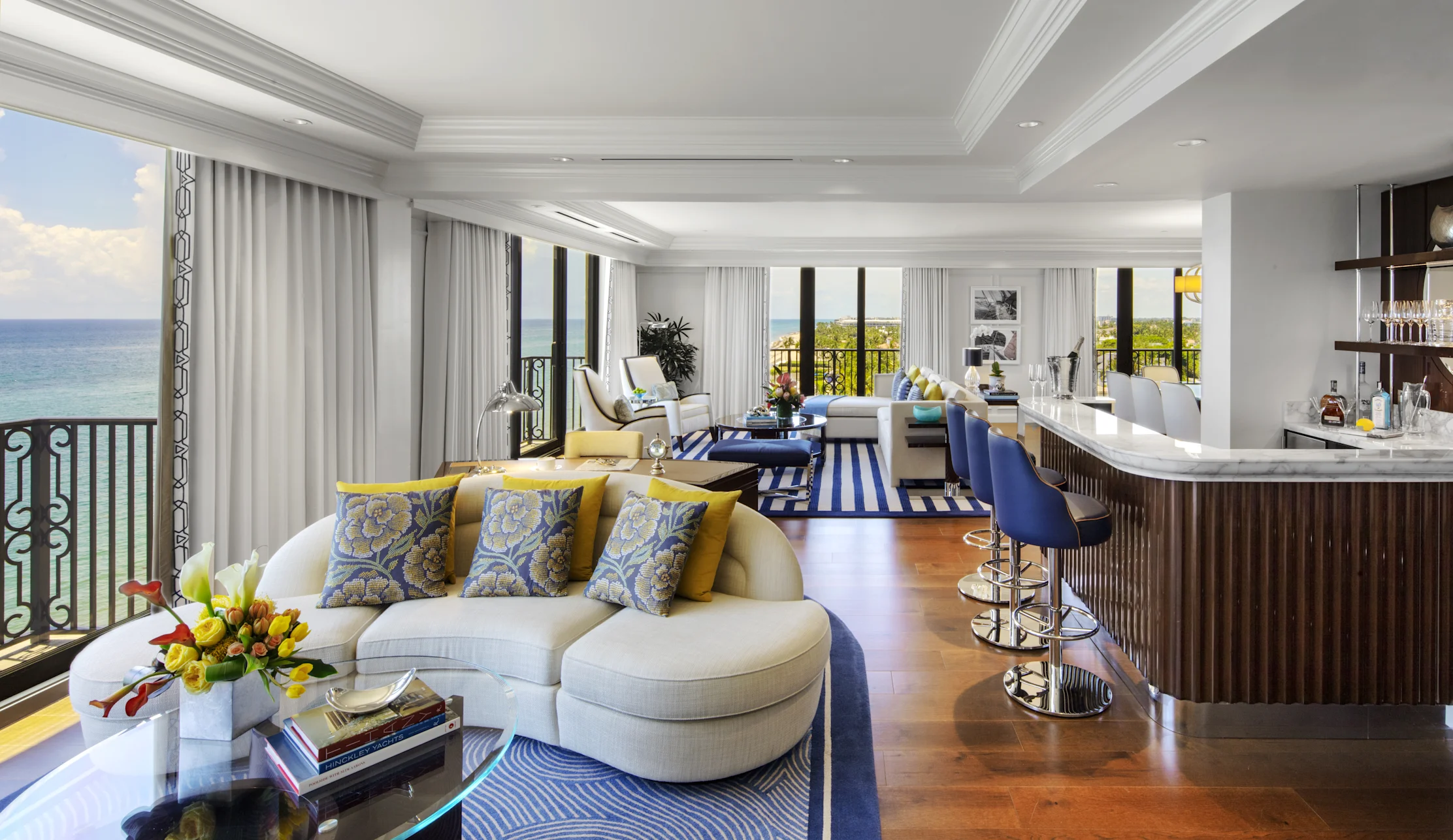 Luxury hotel suite with ocean view, white curved sofa with blue and yellow pillows, bar area, hardwood floors.
