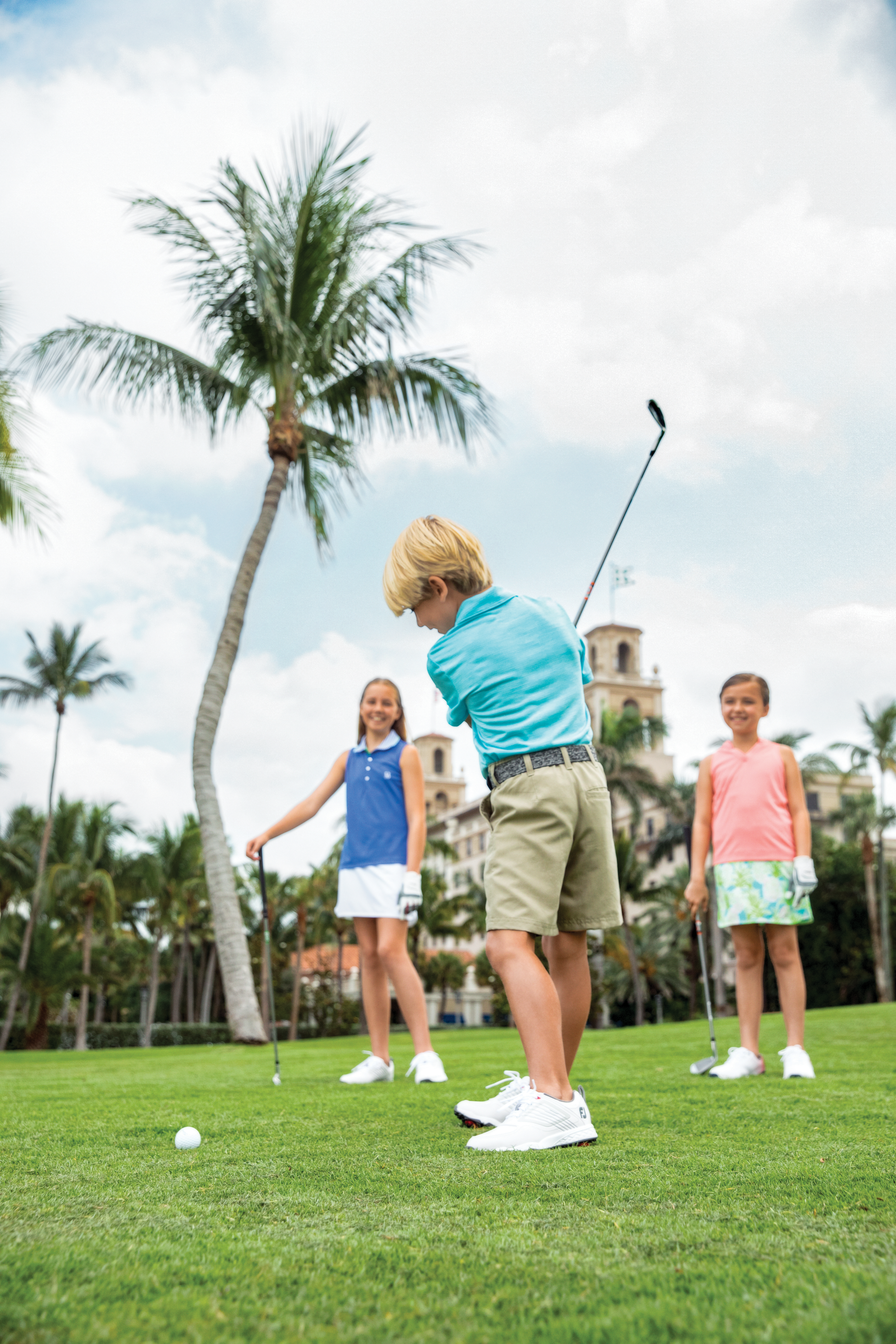 Boy in blue shirt putting golf ball on green lawn with two girls watching and palm trees in background.