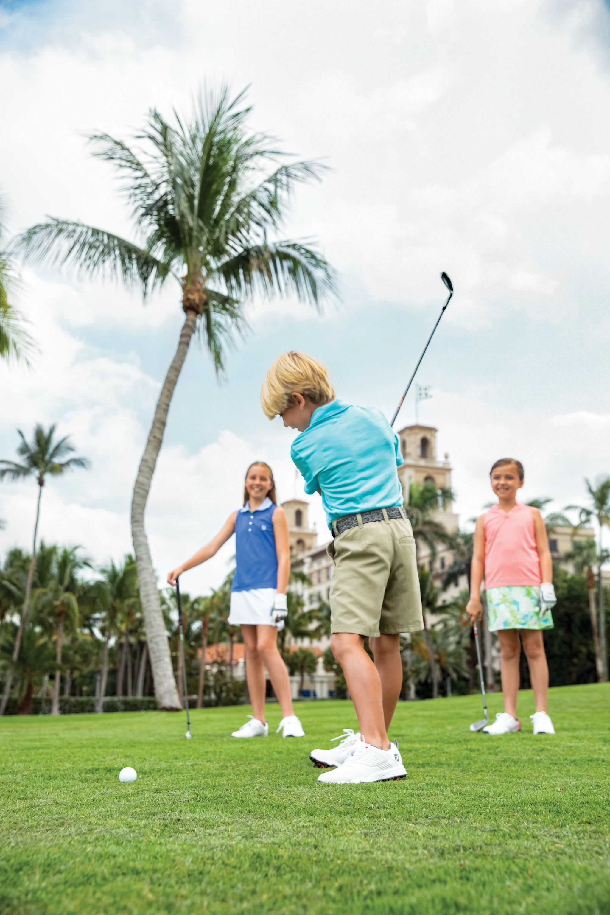 Boy in blue shirt putting golf ball on green lawn with two girls watching and palm trees in background.