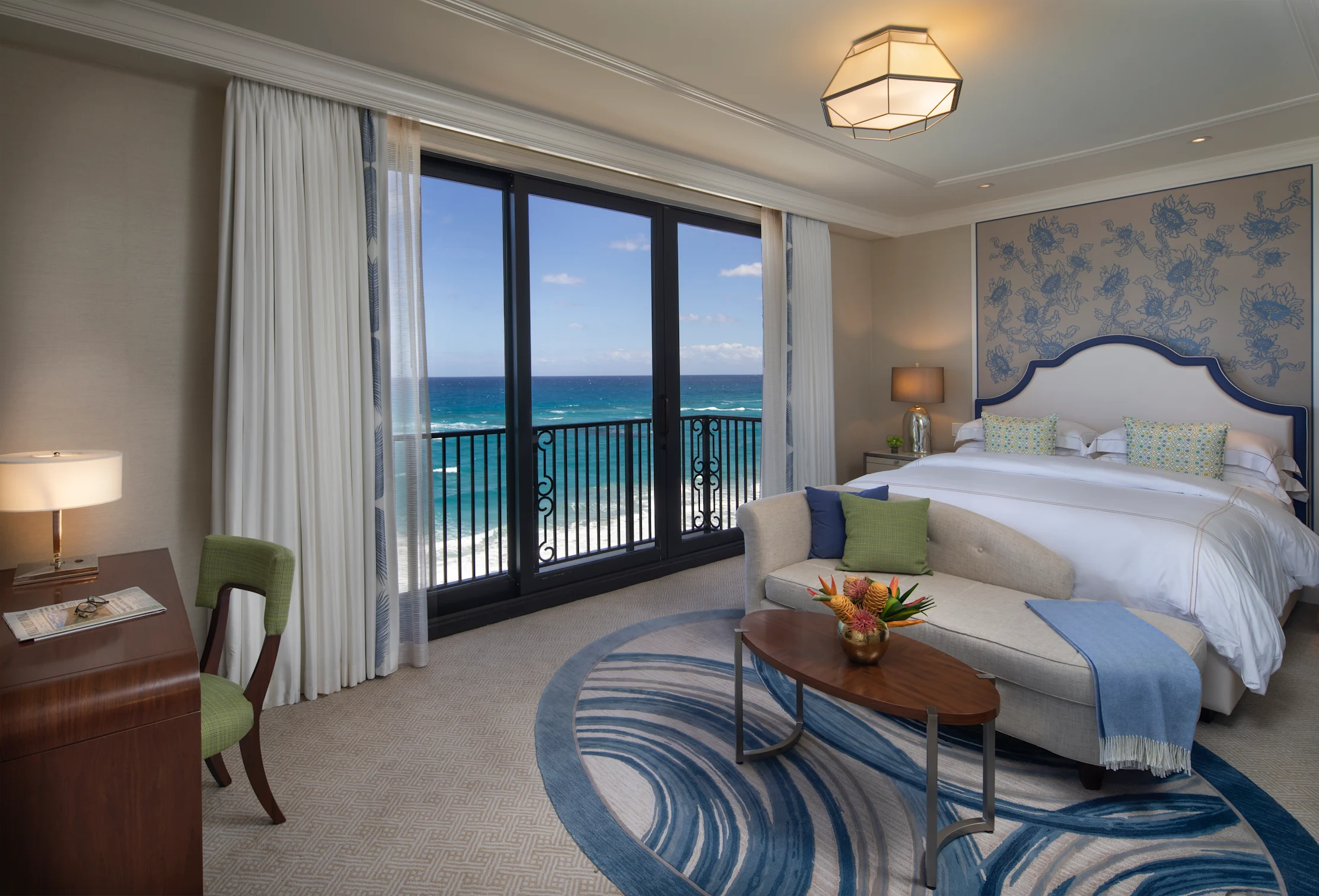Elegant hotel bedroom with ocean view through balcony doors, white bed, beige sofa, and blue accents.