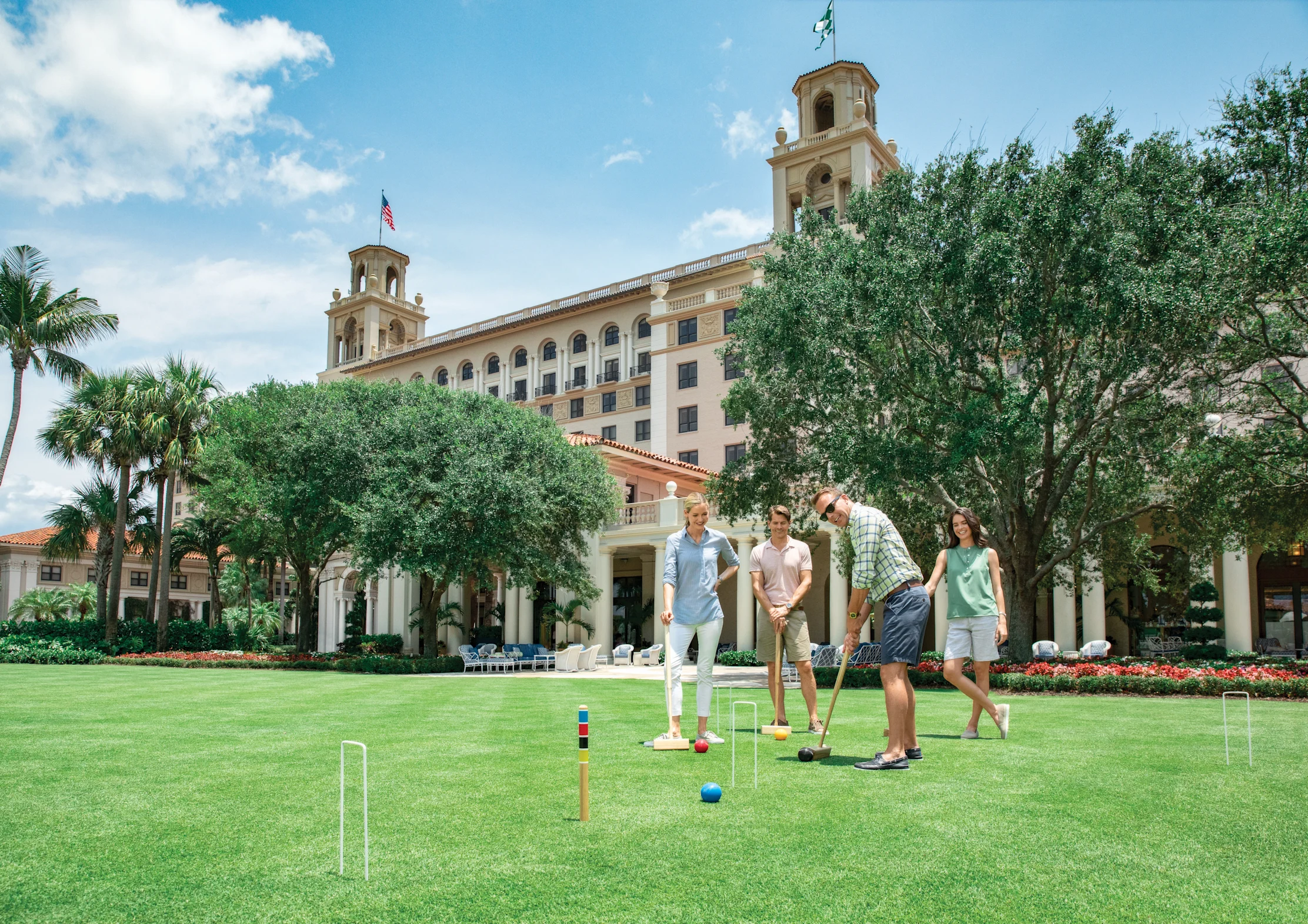 Four people playing croquet on a green lawn in front of a beige Mediterranean-style resort building.