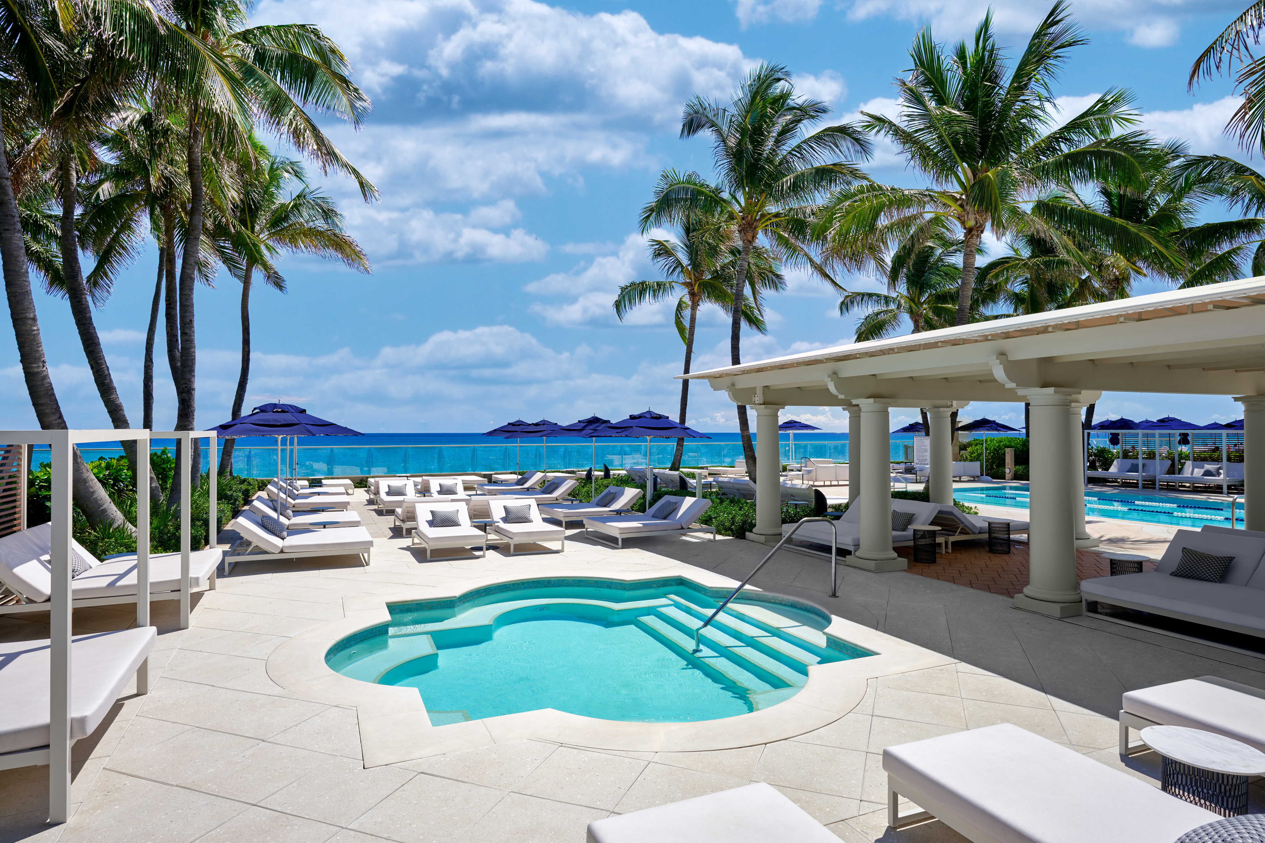 Luxury beachfront resort with turquoise pool, white loungers, palm trees, and ocean view under blue sky.