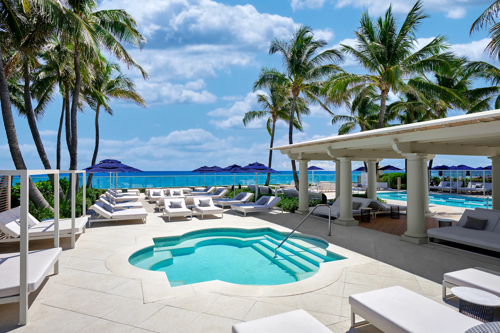Luxury beachfront resort with turquoise pool, white loungers, palm trees, and ocean view under blue sky.