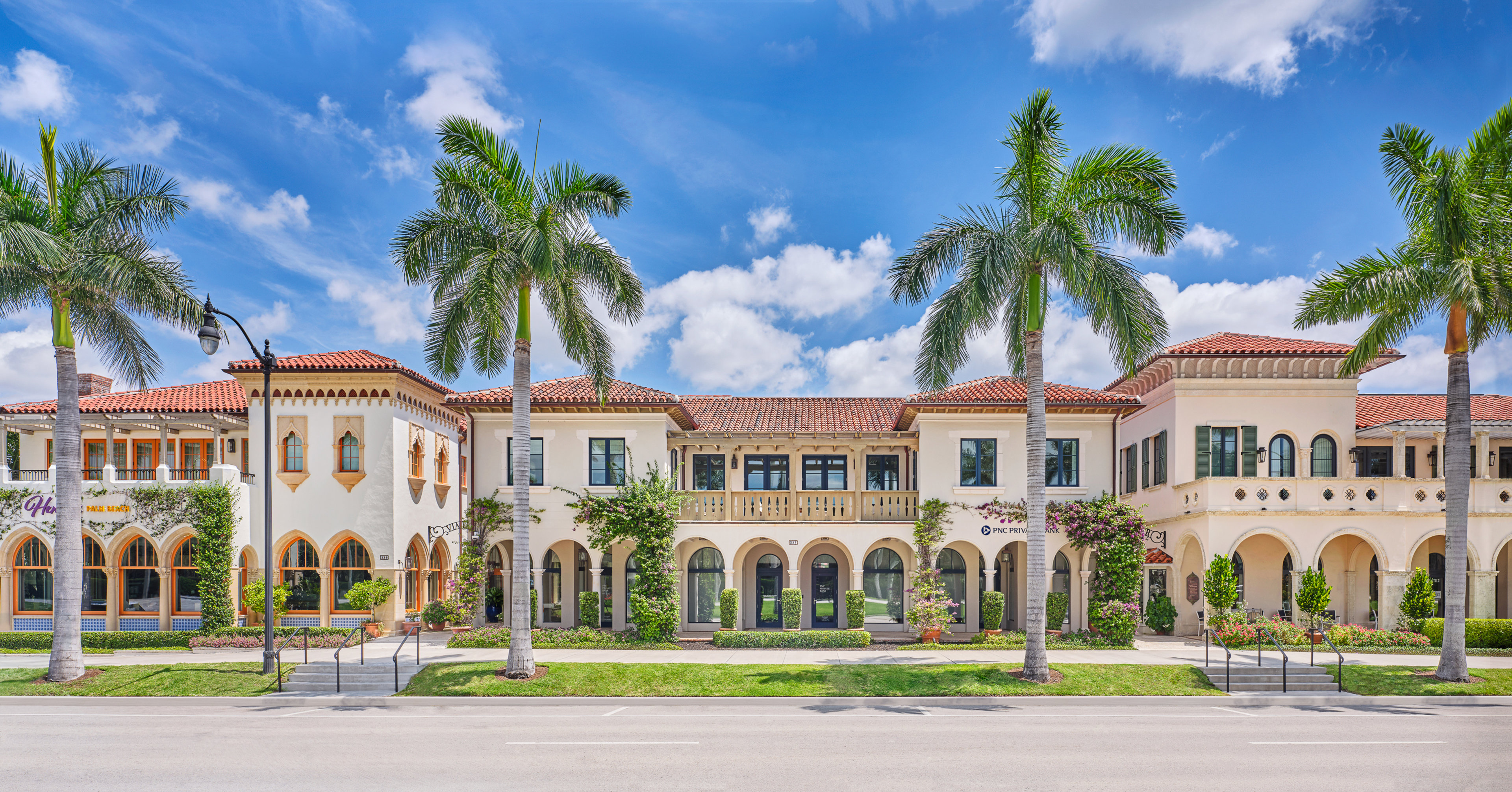 Mediterranean-style commercial buildings with red tile roofs, arched walkways, and palm trees under blue sky.