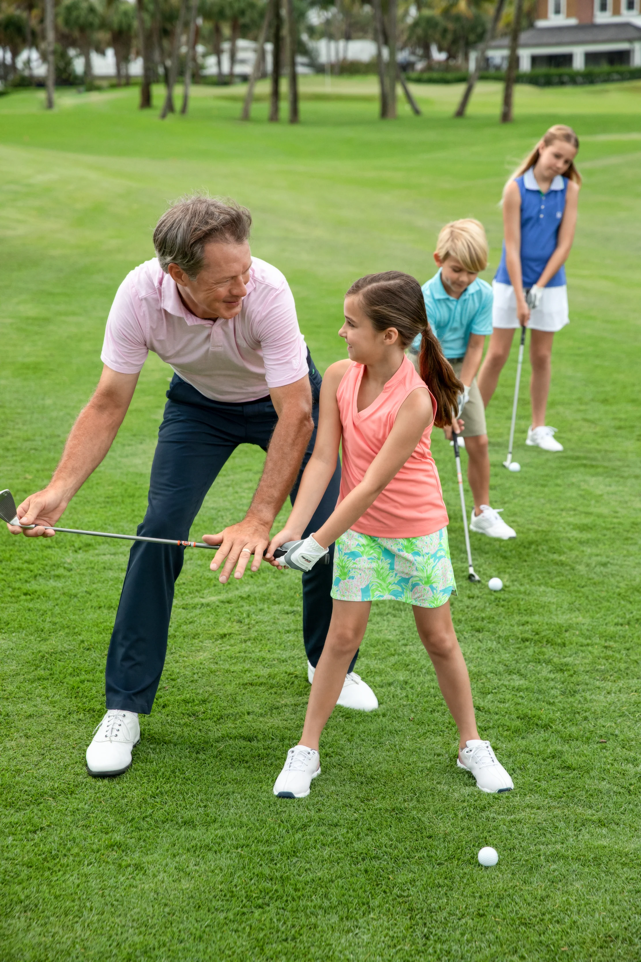 Father teaching daughter to golf on green course with two children watching in background.