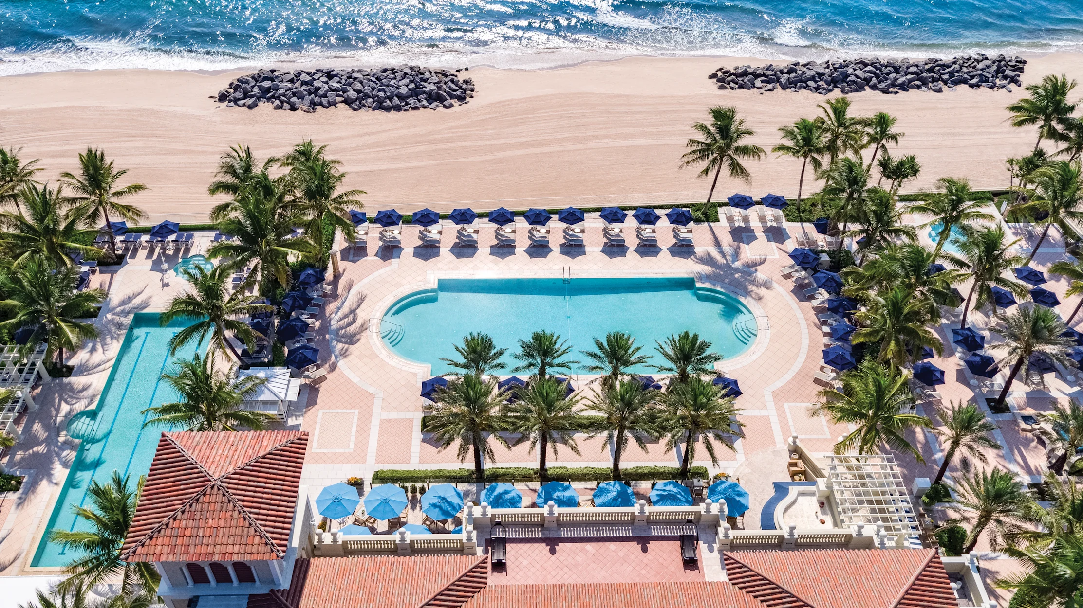 Aerial view of beachfront resort with blue pool, palm trees, lounge chairs, and sandy beach.