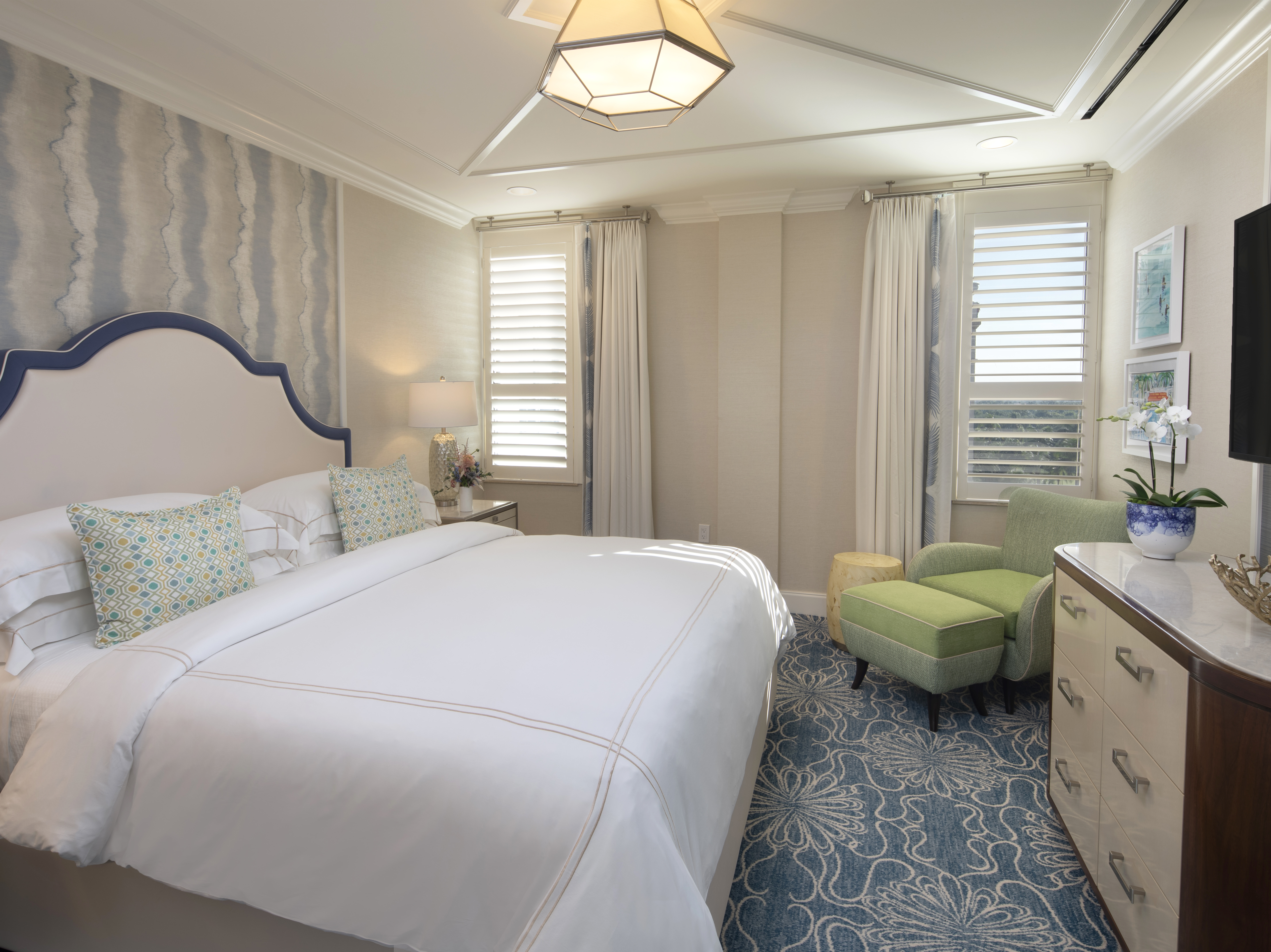Elegant bedroom with white upholstered bed, geometric pendant light, green armchair, and blue patterned rug.