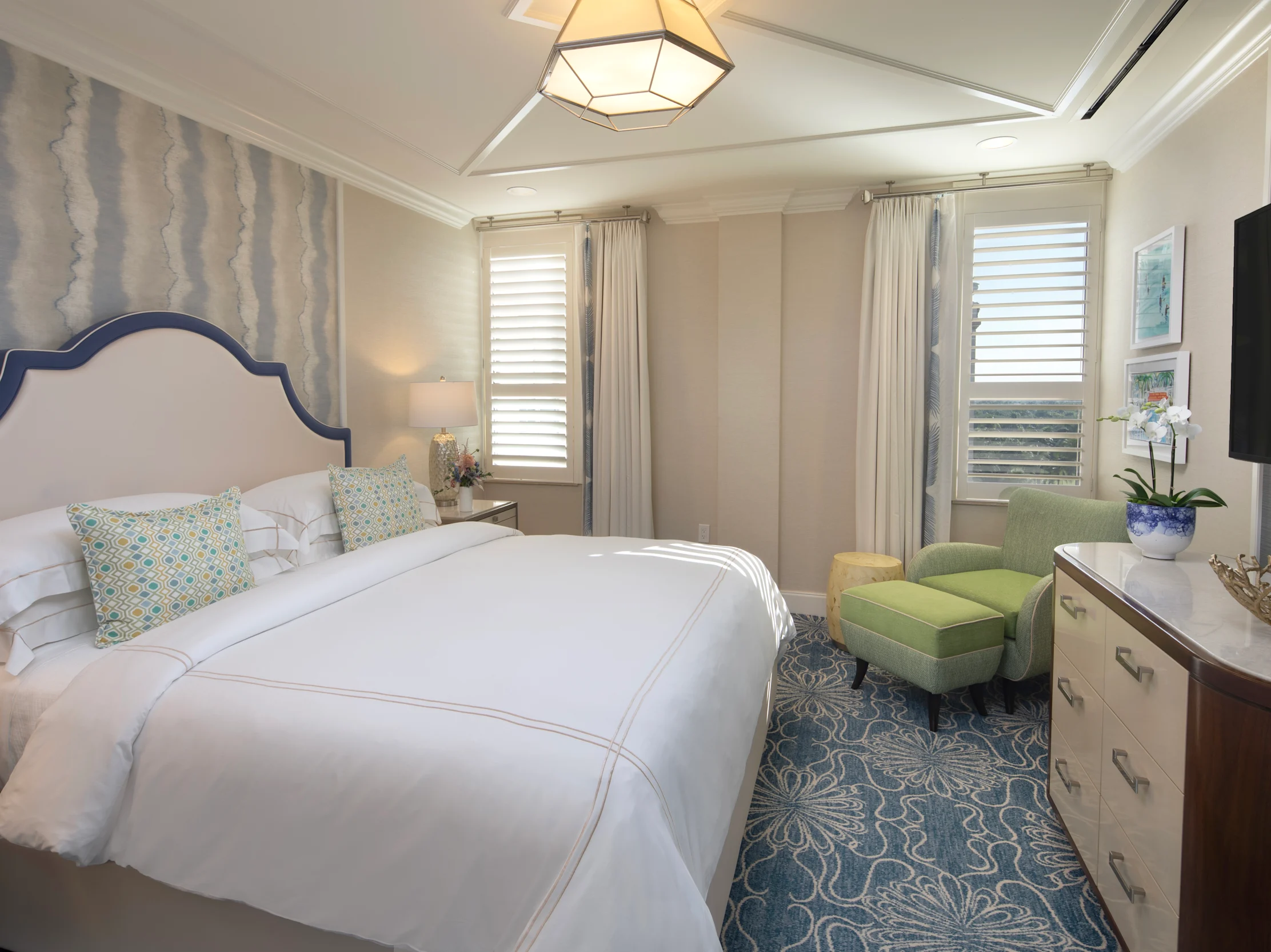 Elegant bedroom with white upholstered bed, geometric pendant light, green armchair, and blue patterned rug.