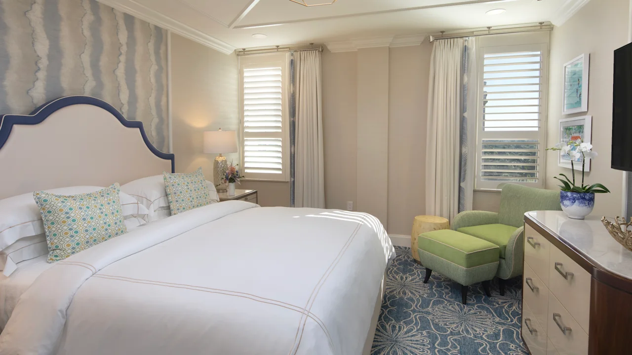 Elegant bedroom with white upholstered bed, geometric pendant light, green armchair, and blue patterned rug.