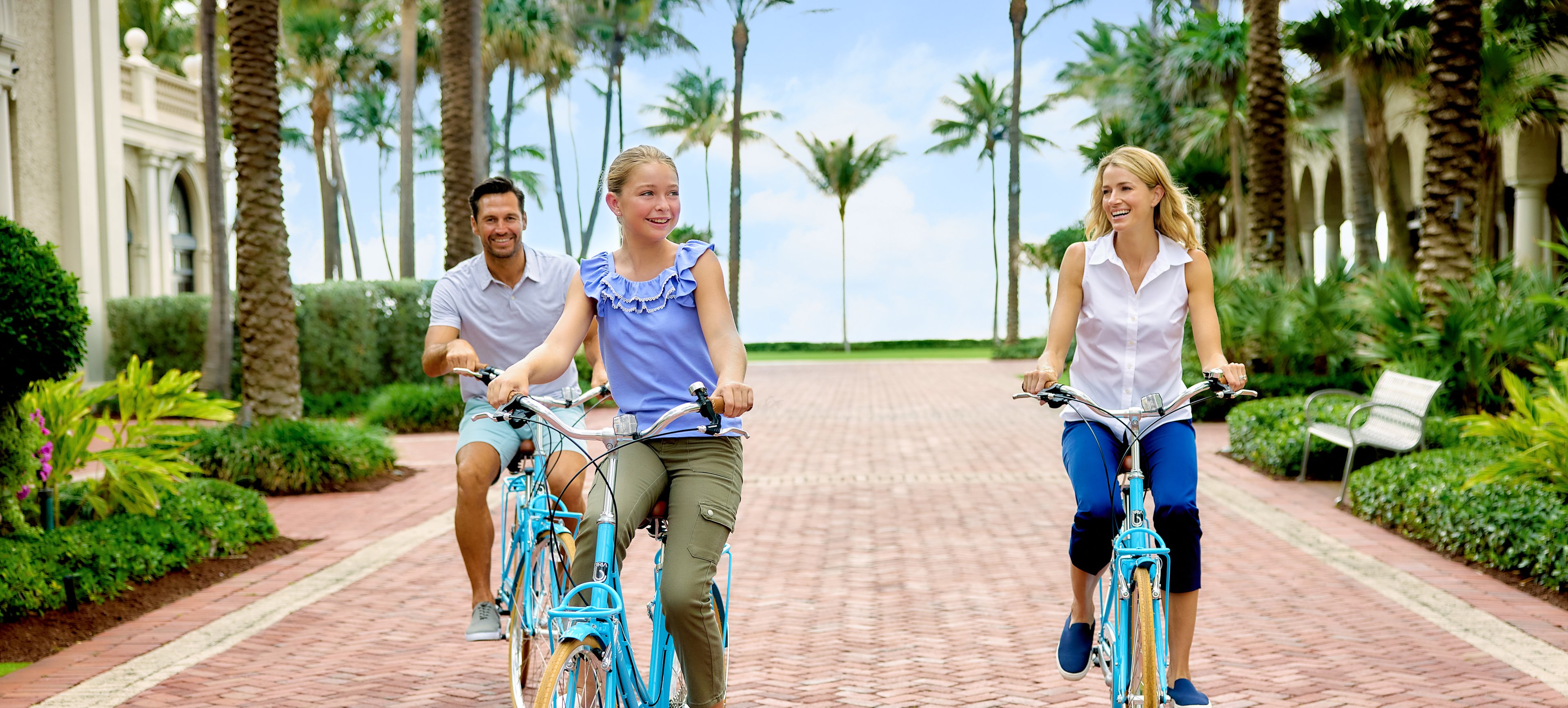 Family of three riding blue bicycles on brick path lined with palm trees at tropical resort.