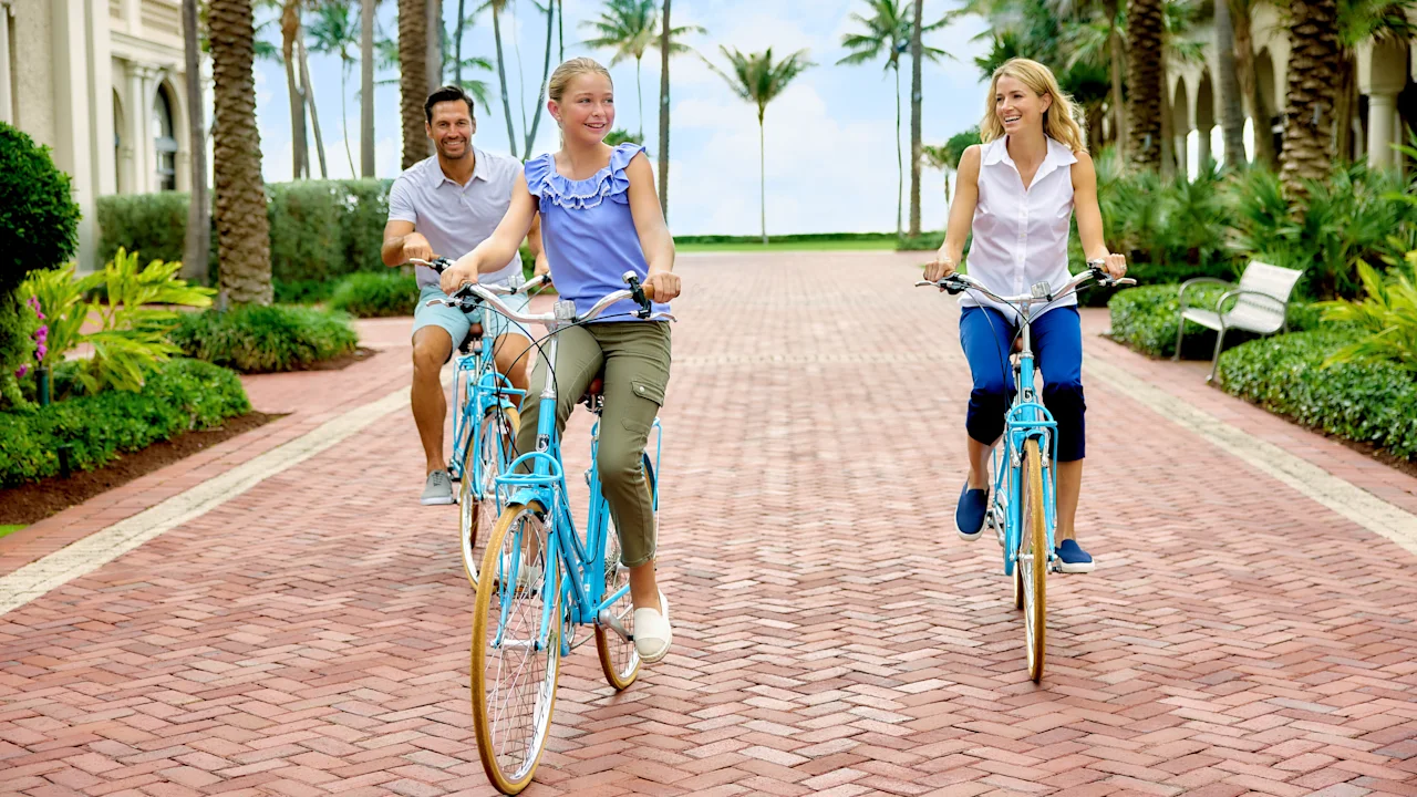 Family of three riding blue bicycles on brick path lined with palm trees at tropical resort.