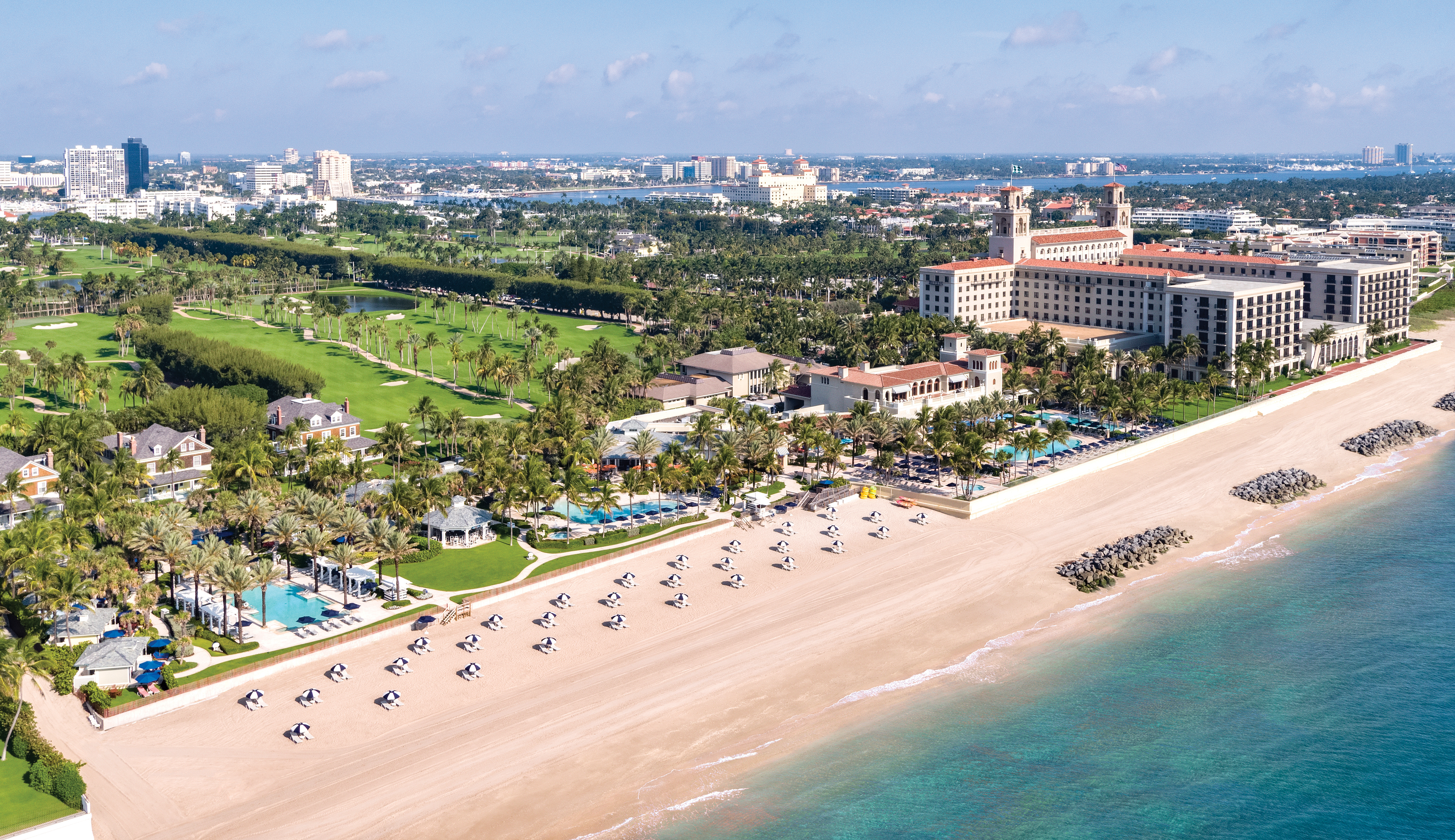 Aerial view of beachfront resort with white sand, turquoise water, palm trees, and golf course.
