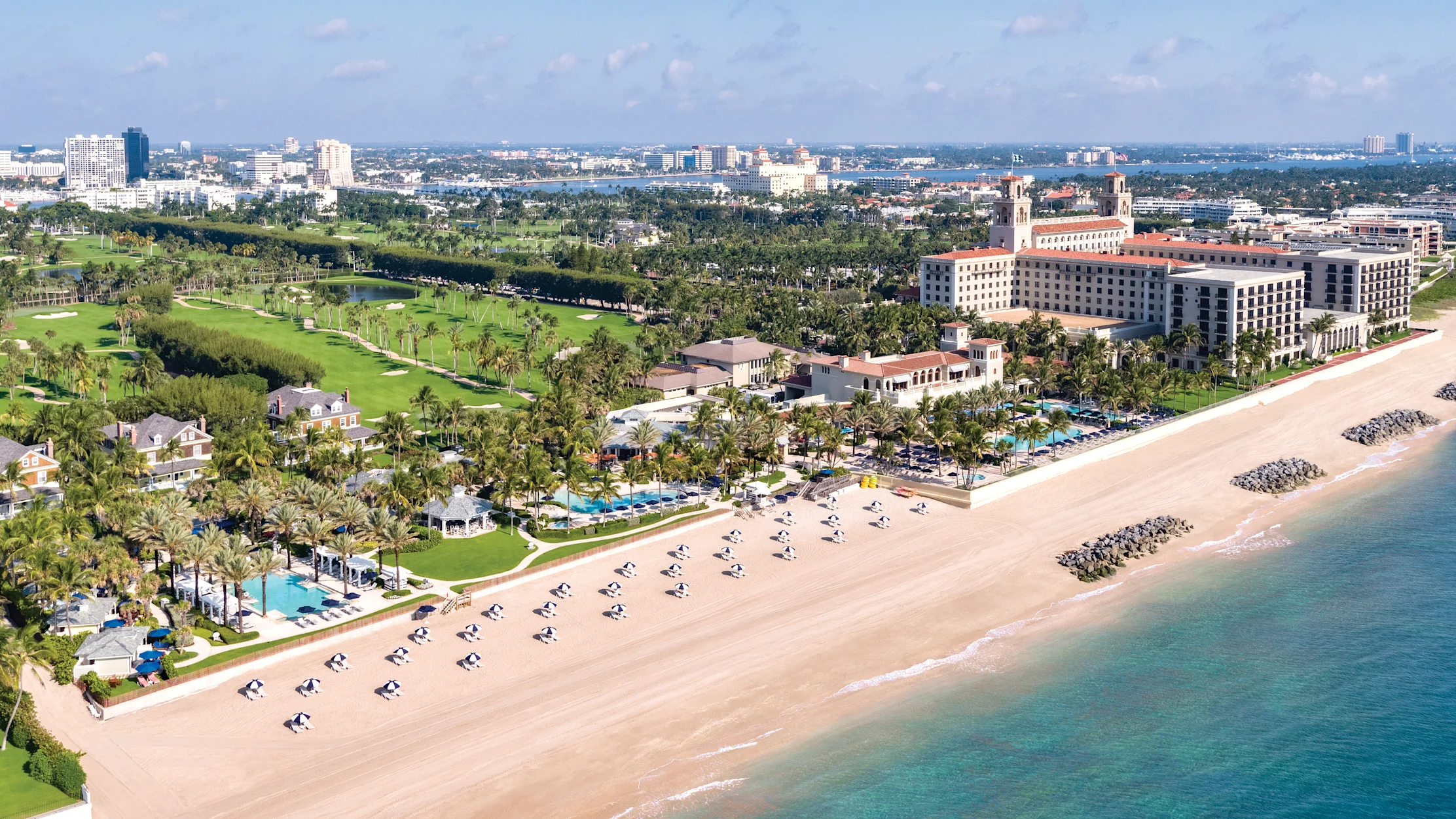 Aerial view of beachfront resort with white sand, turquoise water, palm trees, and golf course.