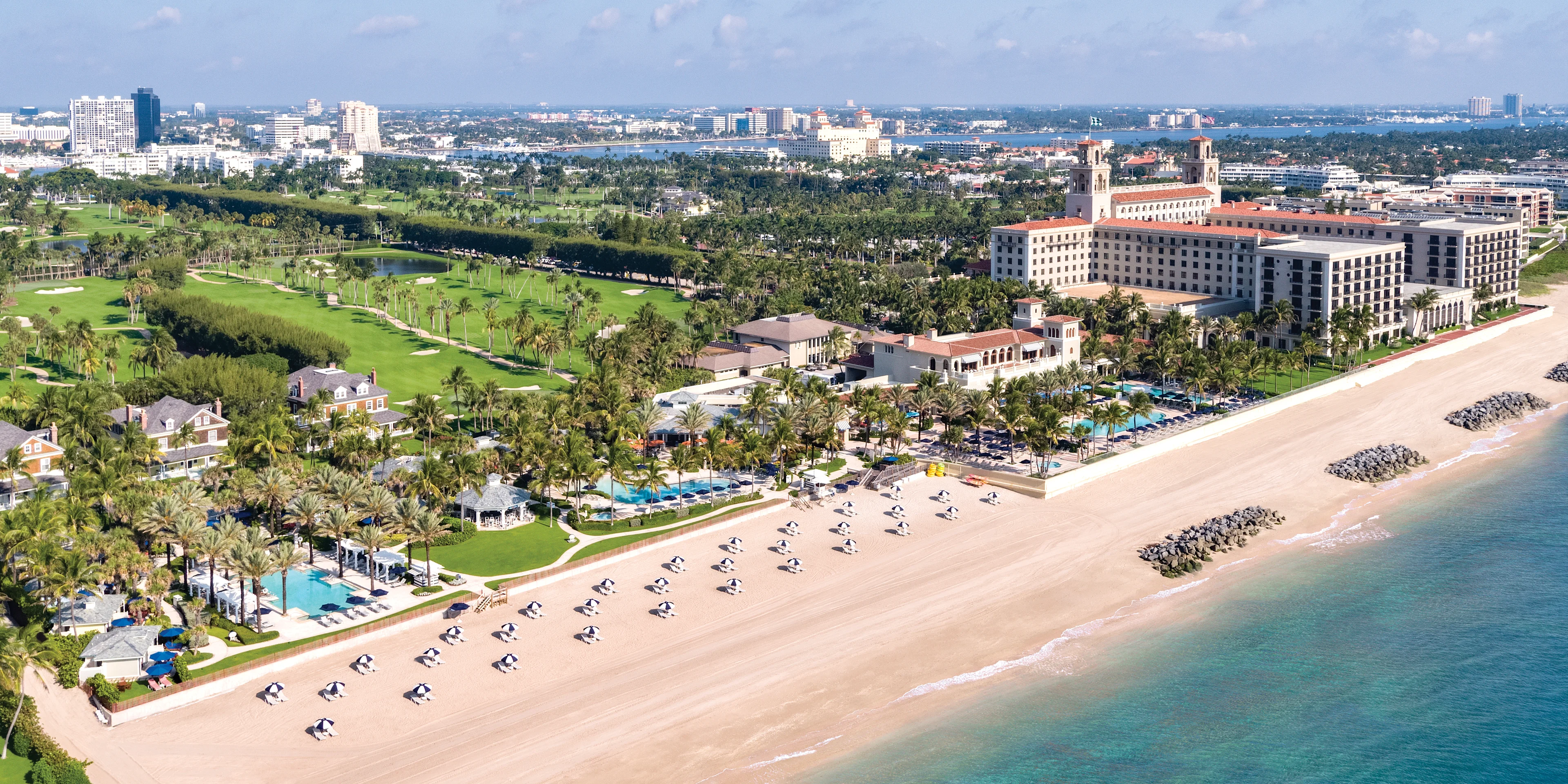 Aerial view of beachfront resort with white sand, turquoise water, palm trees, and golf course.