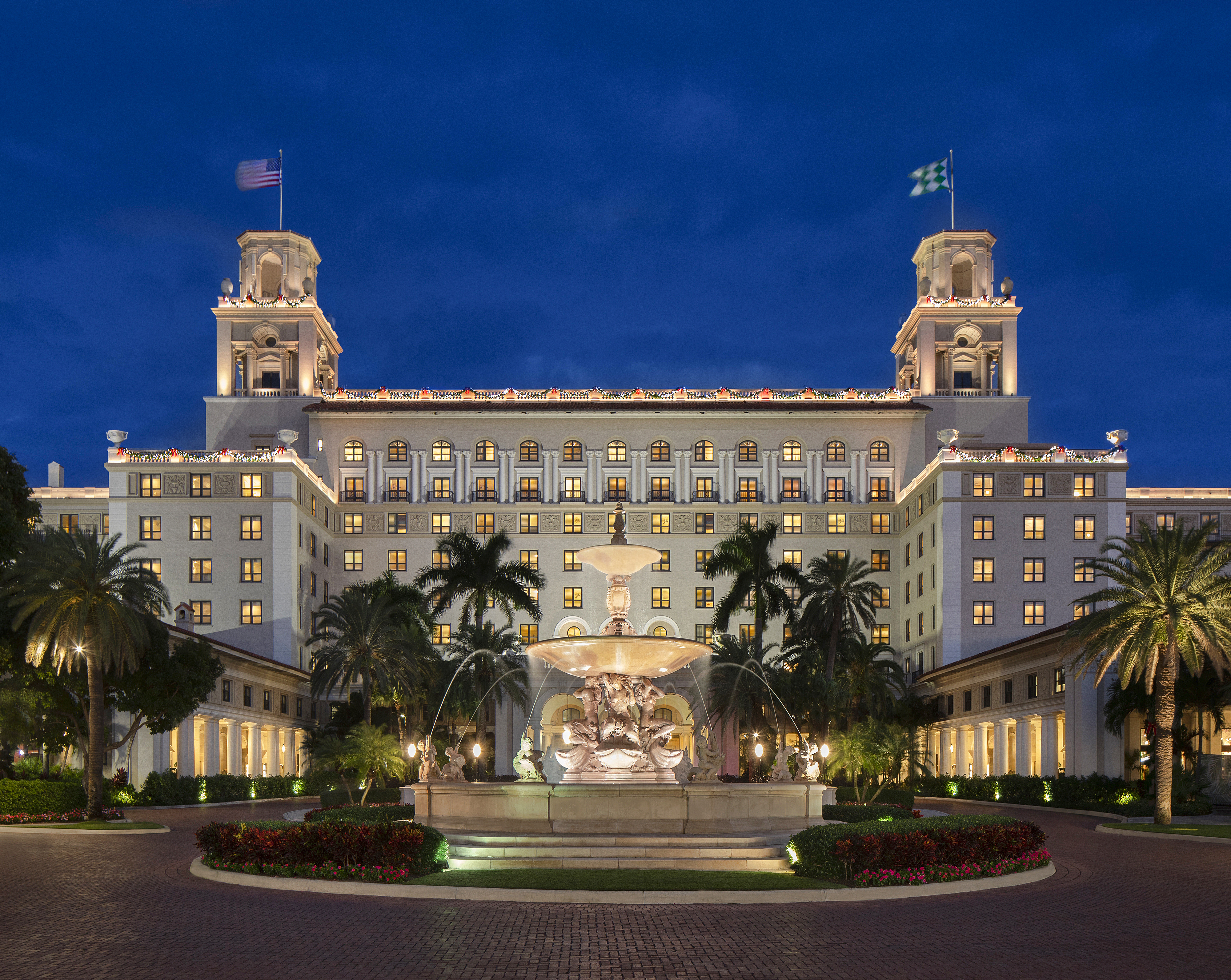 Illuminated white hotel with twin towers and fountain at dusk, surrounded by palm trees.
