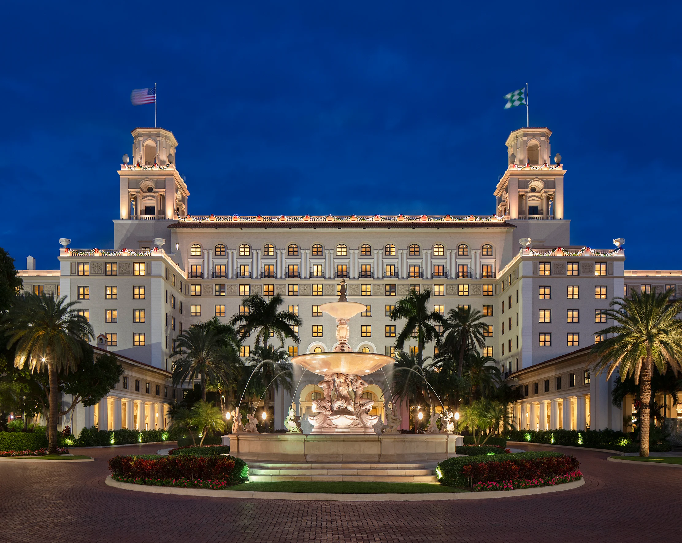 Illuminated white hotel with twin towers and fountain at dusk, surrounded by palm trees.