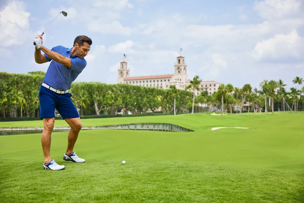Golfer in blue striped shirt swinging club on green course with resort building in background.