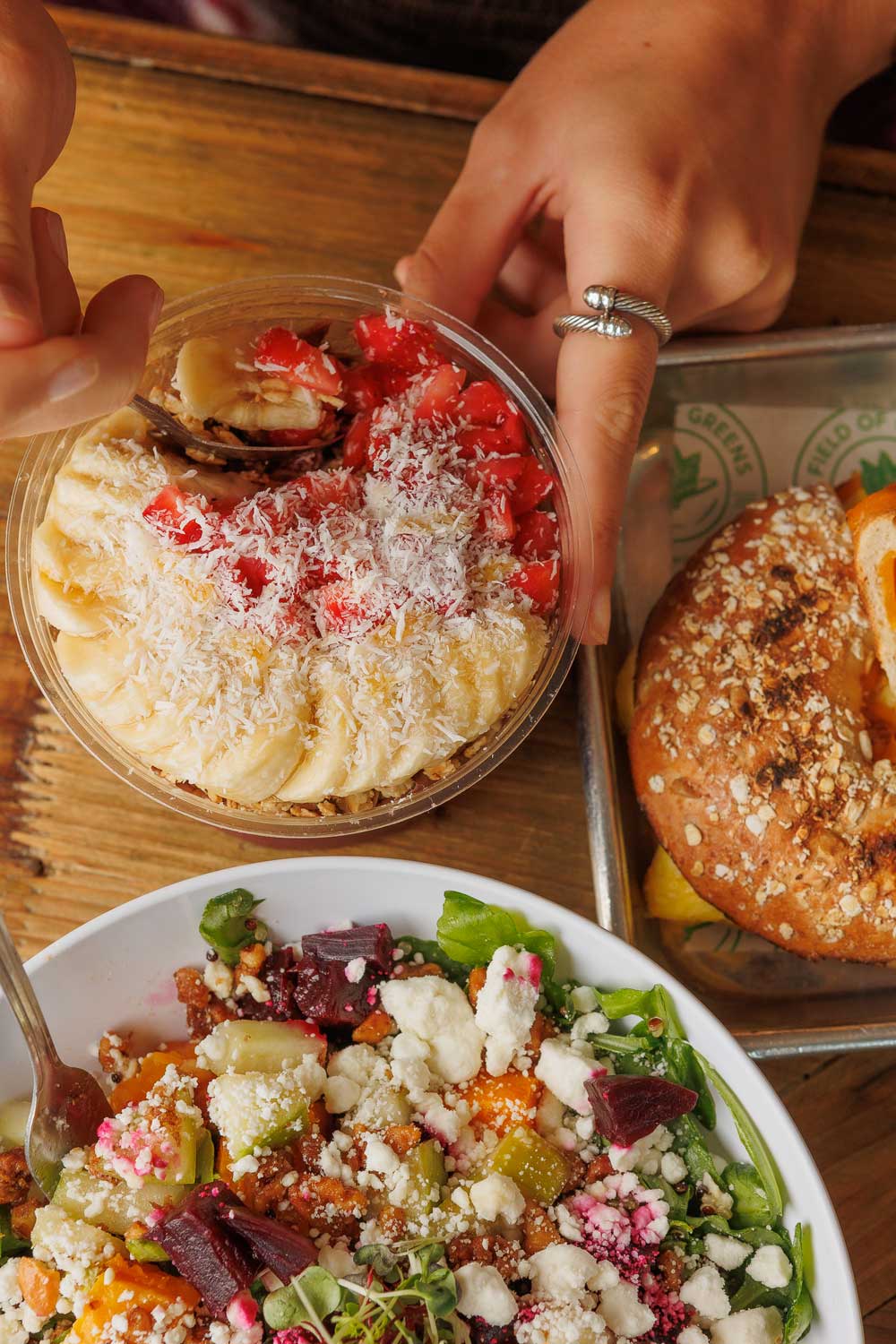 Hand reaching for acai bowl topped with bananas, strawberries, and coconut next to salad and pretzel