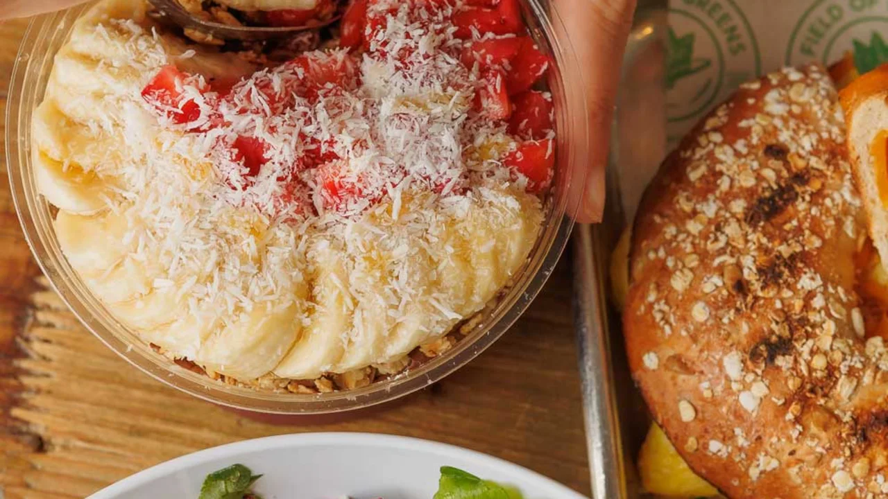 Hand reaching for acai bowl topped with bananas, strawberries, and coconut next to salad and pretzel