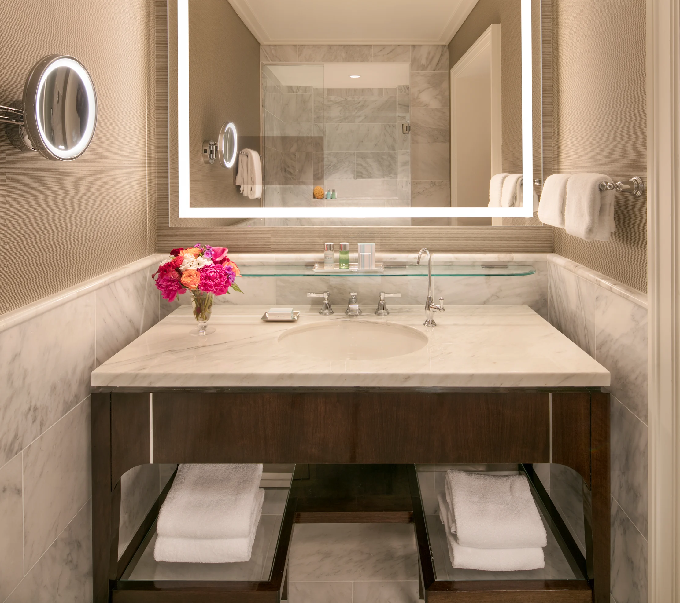 Modern hotel bathroom with marble vanity, LED-lit mirror, pink flowers, and beige tile walls.
