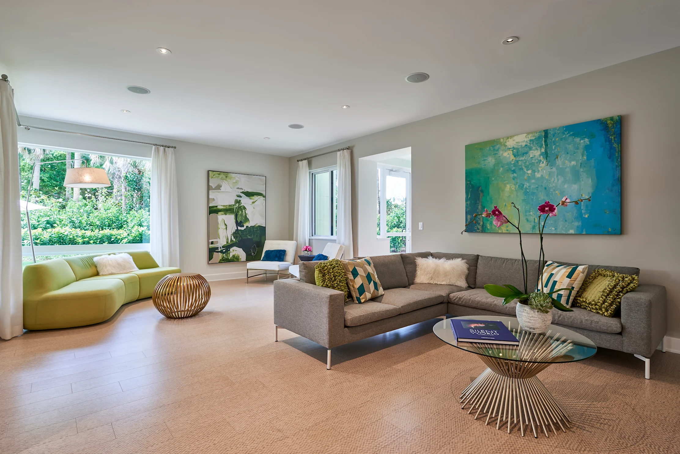 Modern living room with gray sofa, lime green curved couch, abstract blue art, and garden views.