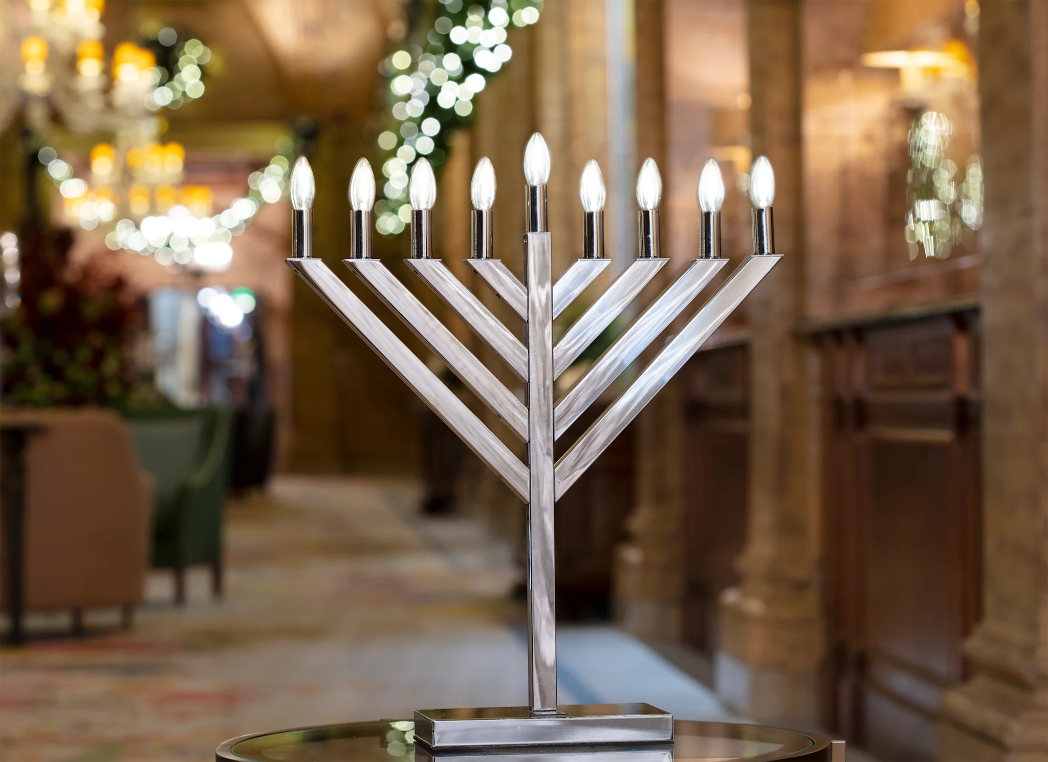 Silver menorah with nine lit candles displayed in a warmly decorated room with holiday lights.