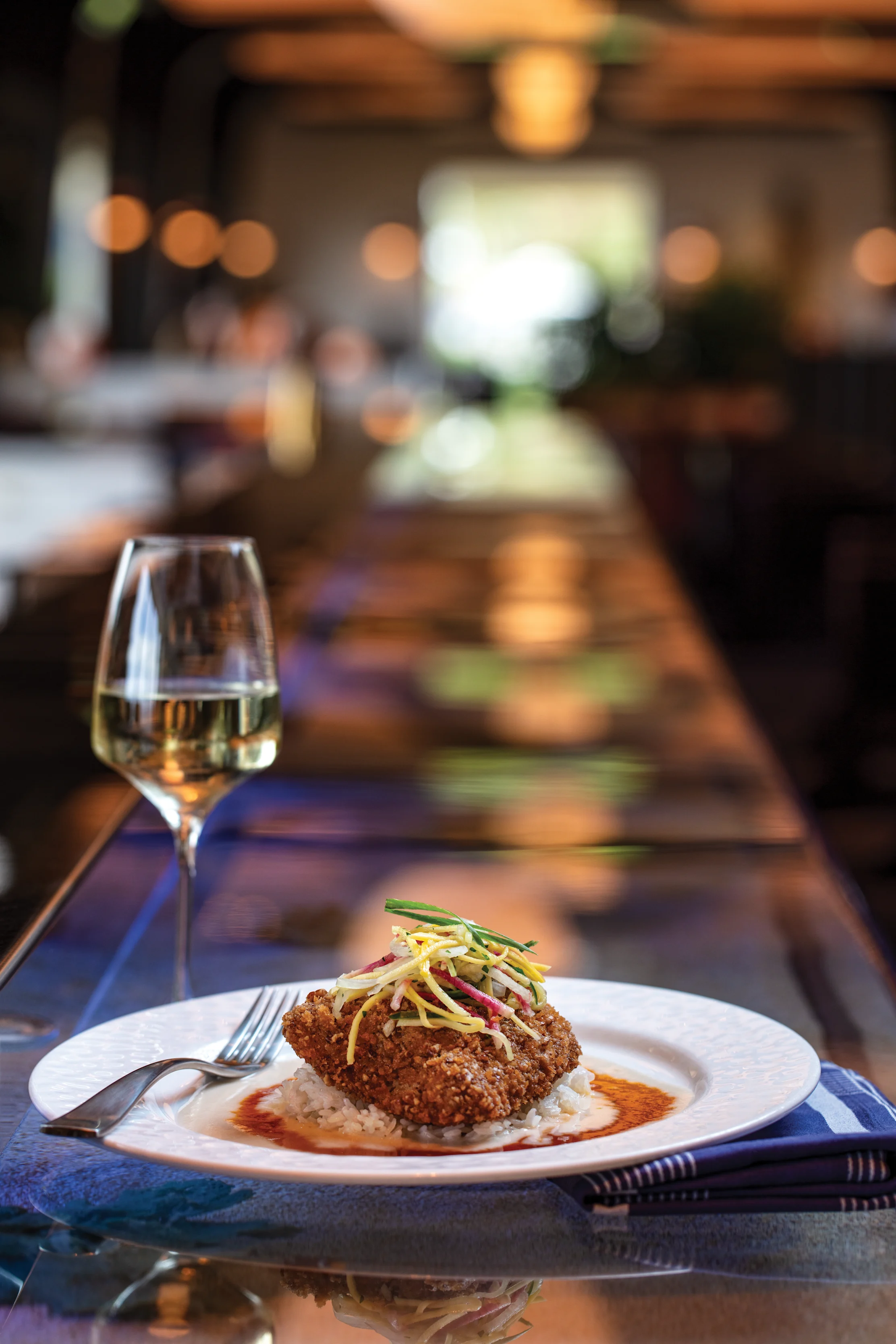 Breaded cutlet with slaw on white plate beside white wine glass in warm restaurant setting