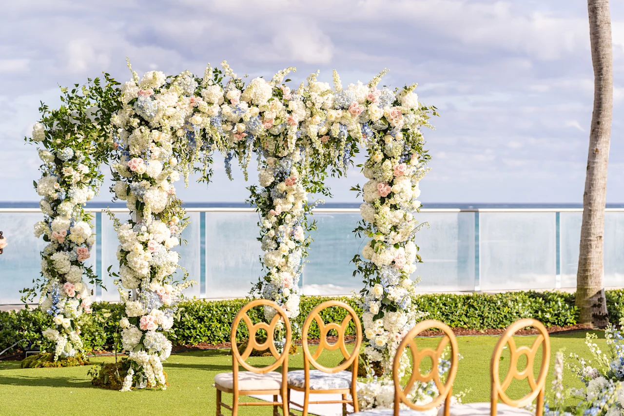 Four-post ceremony chuppah covered in white hydrangea and blush roses on an oceanfront lawn, with gold ring-back chairs and the sea beyond a glass railing.