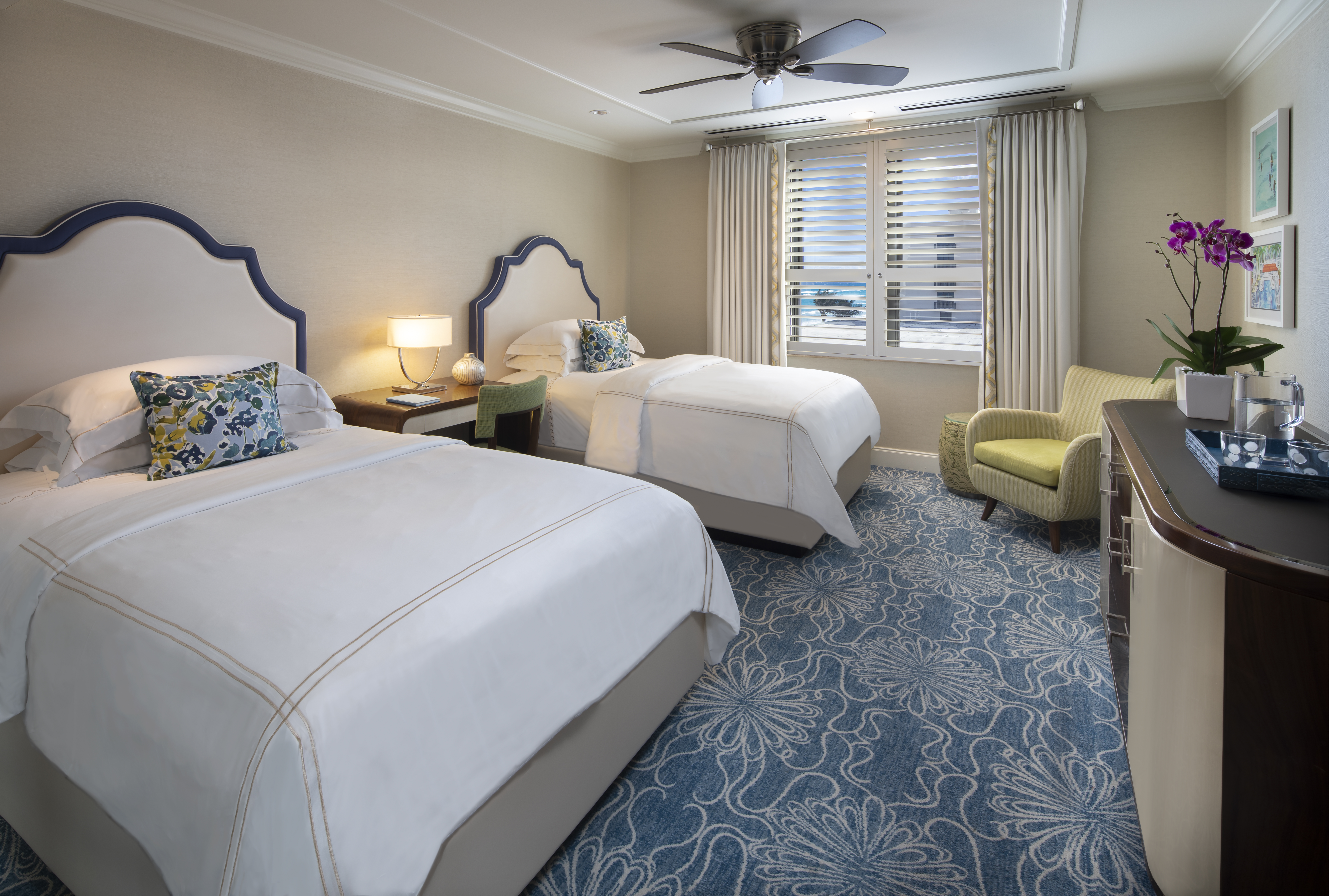 Elegant hotel room with two beds featuring navy headboards, white linens, blue patterned carpet, and yellow accent chair.