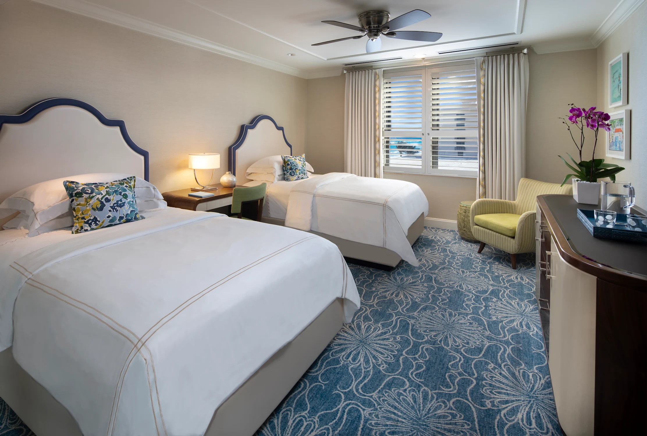 Elegant hotel room with two beds featuring navy headboards, white linens, blue patterned carpet, and yellow accent chair.