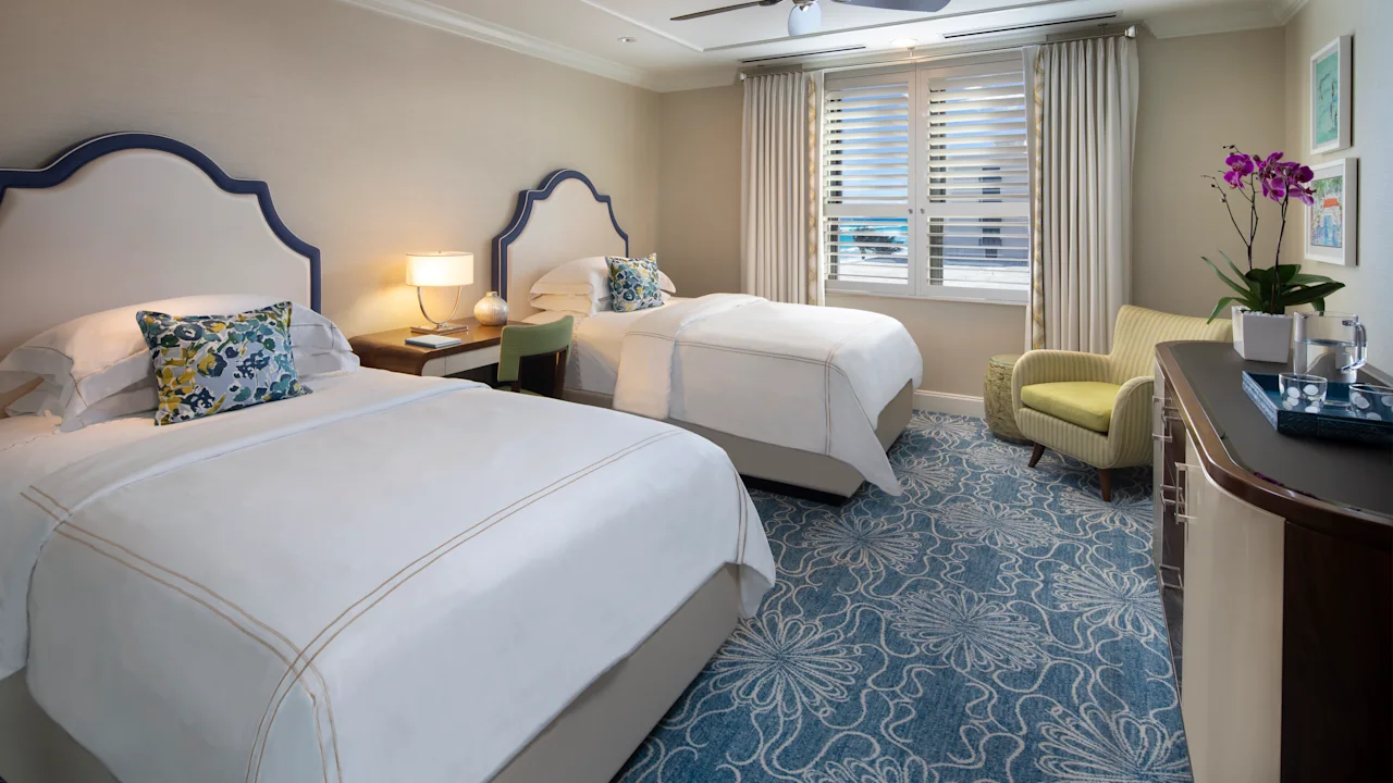 Elegant hotel room with two beds featuring navy headboards, white linens, blue patterned carpet, and yellow accent chair.