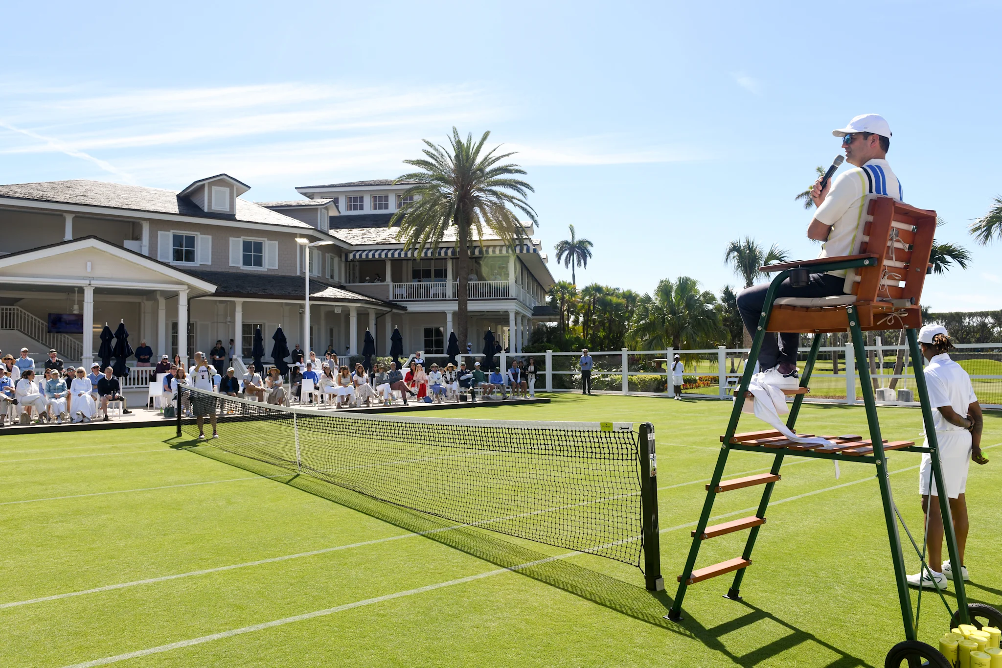 Tennis match at upscale club with umpire on chair, spectators, and white clubhouse with palm trees.