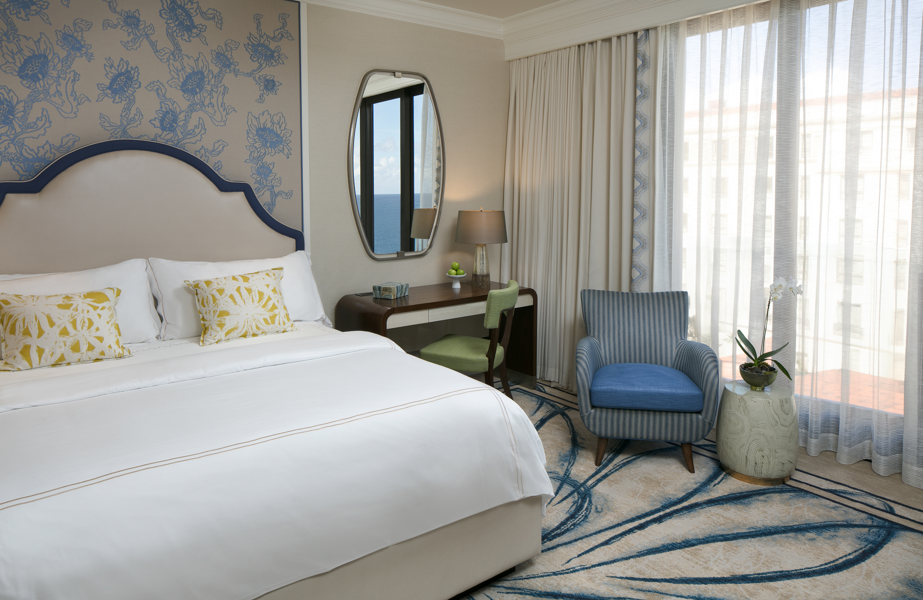 Elegant hotel bedroom with white bed, blue patterned wallpaper, oval mirror, desk, and blue armchair.