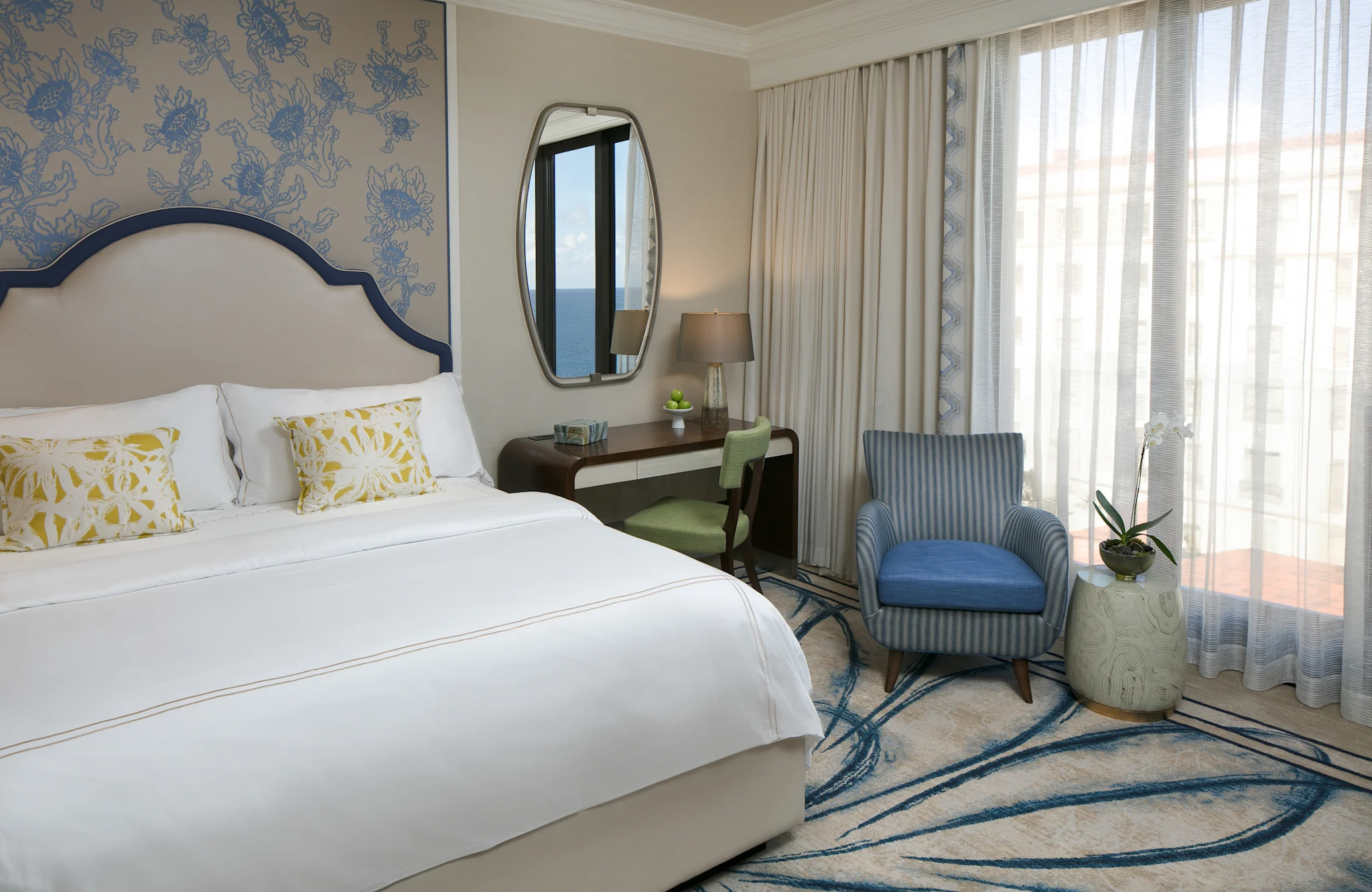 Elegant hotel bedroom with white bed, blue patterned wallpaper, oval mirror, desk, and blue armchair.