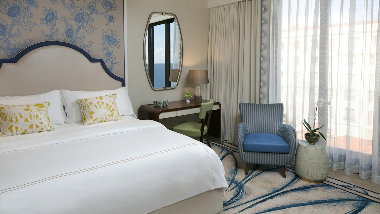 Elegant hotel bedroom with white bed, blue patterned wallpaper, oval mirror, desk, and blue armchair.