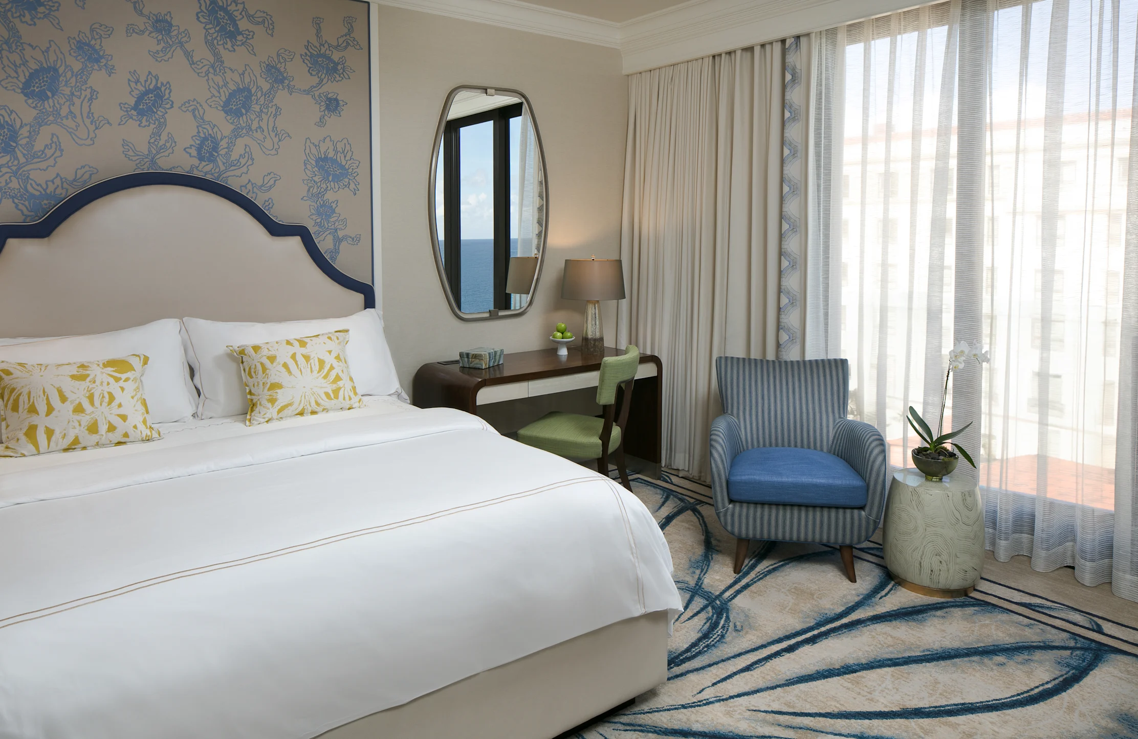Elegant hotel bedroom with white bed, blue patterned wallpaper, oval mirror, desk, and blue armchair.