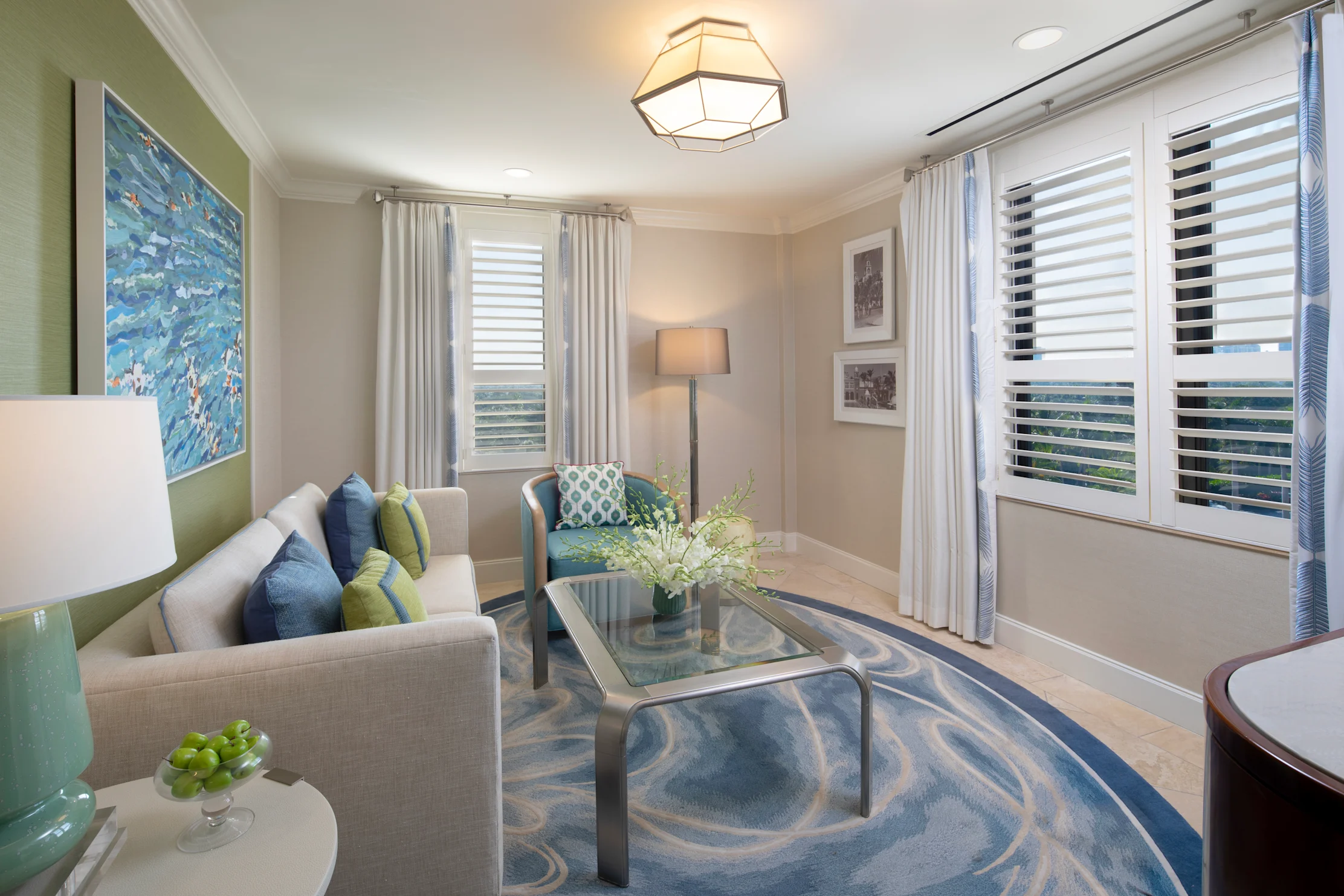 Modern living room with beige sofa, blue and green accents, geometric light fixture, and windows.