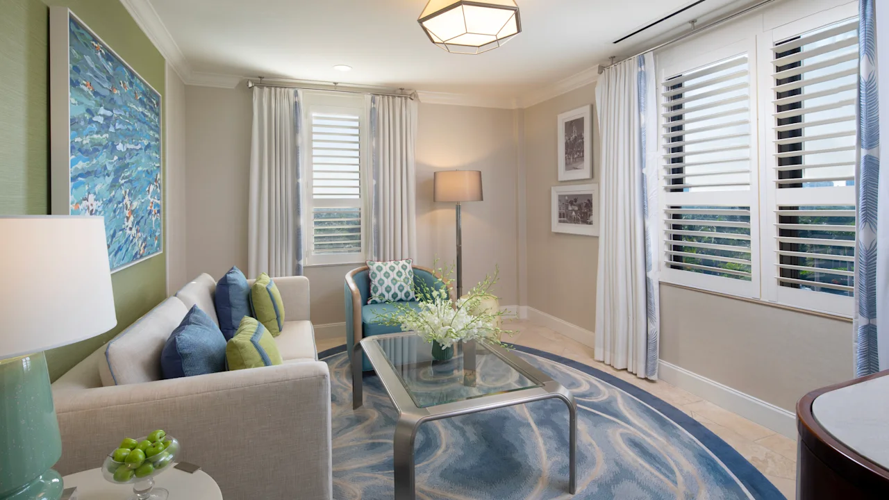 Bright living room with beige sofa, blue and green pillows, glass coffee table, and white shutters.